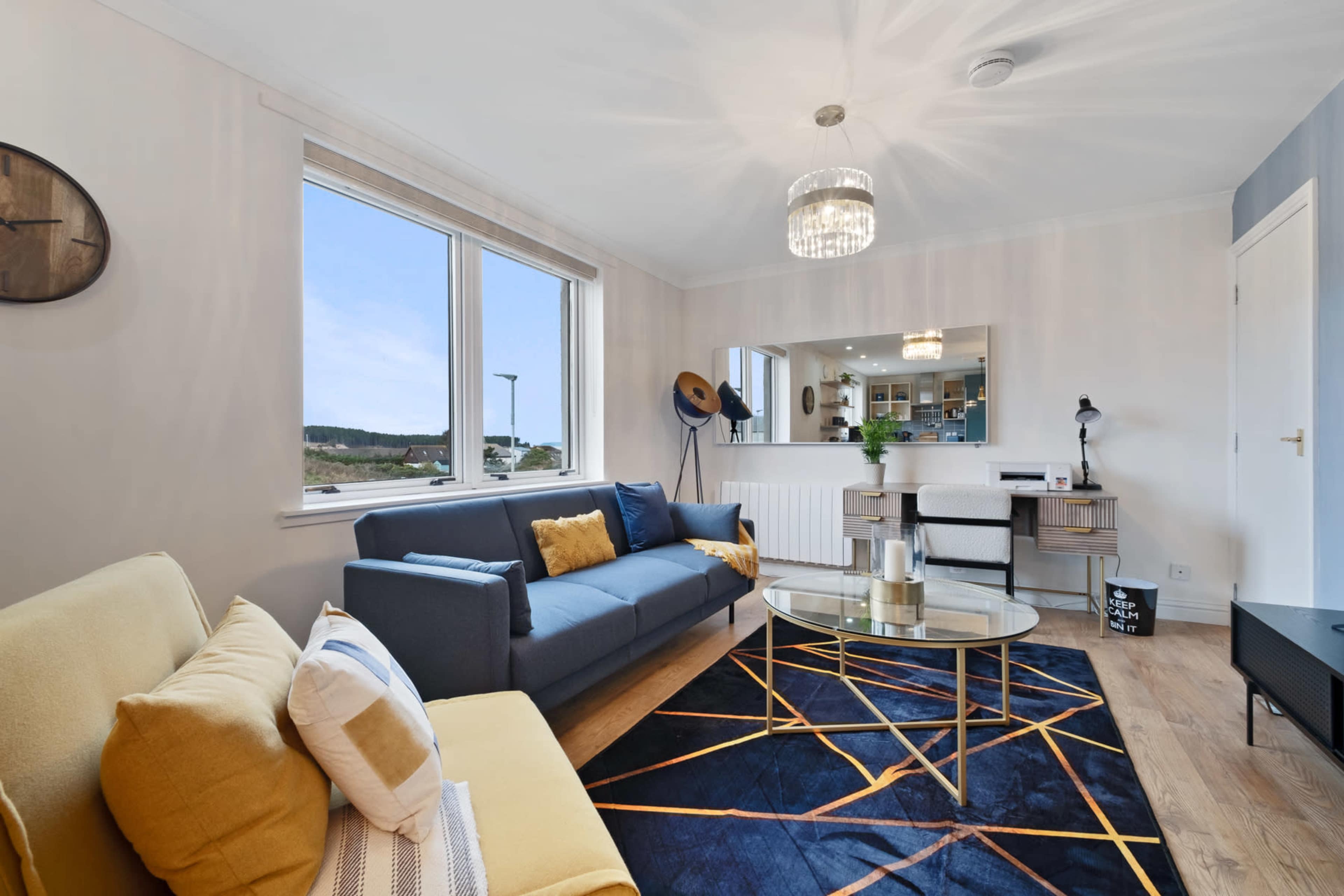 A modern living room with a yellow sofa, a blue couch, a glass coffee table, and large windows allowing natural light to fill the space.