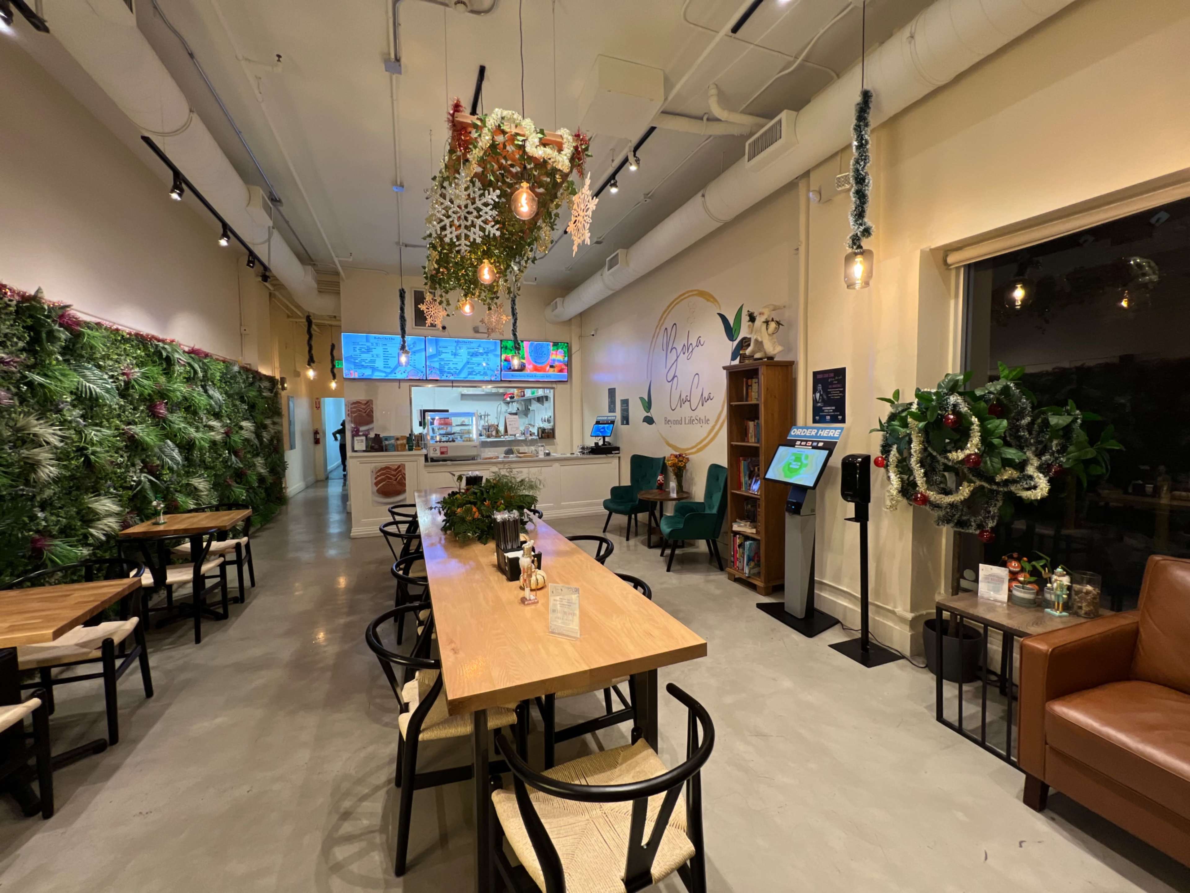 A cafe interior with a row of tables and chairs, green wall decorations, a service counter in the background, and festive plants overhead.