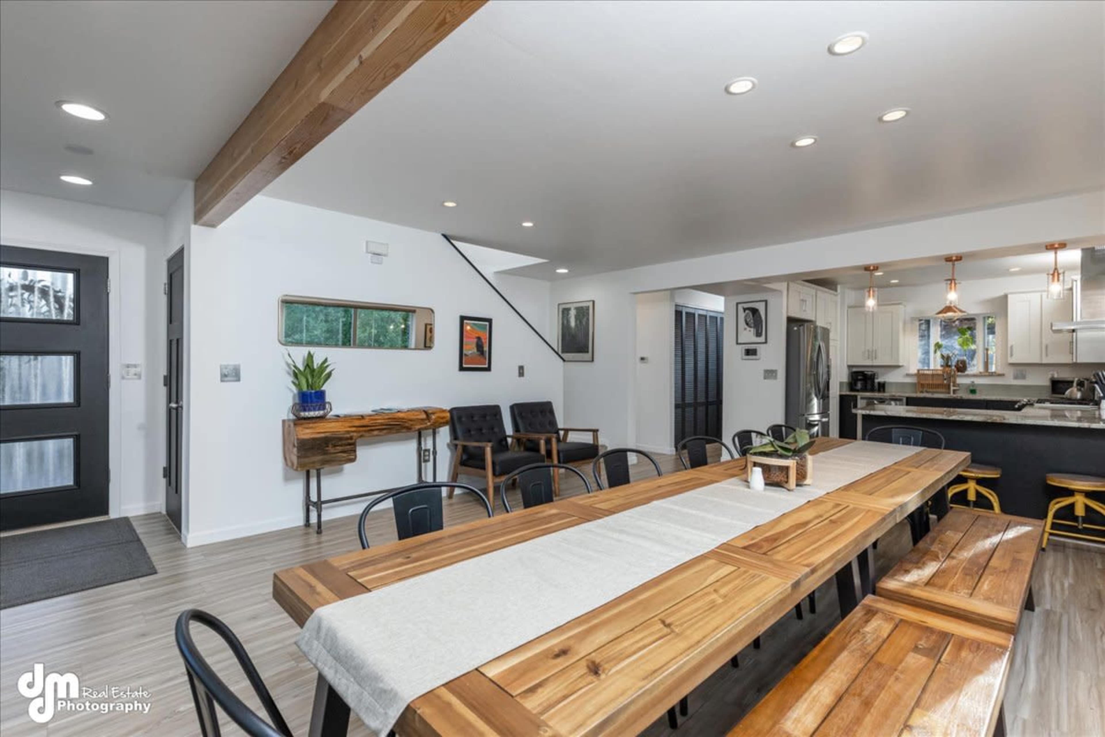 The image shows a modern dining area with a long wooden table surrounded by black chairs, leading to a kitchen in the background with white cabinetry and stainless steel appliances.