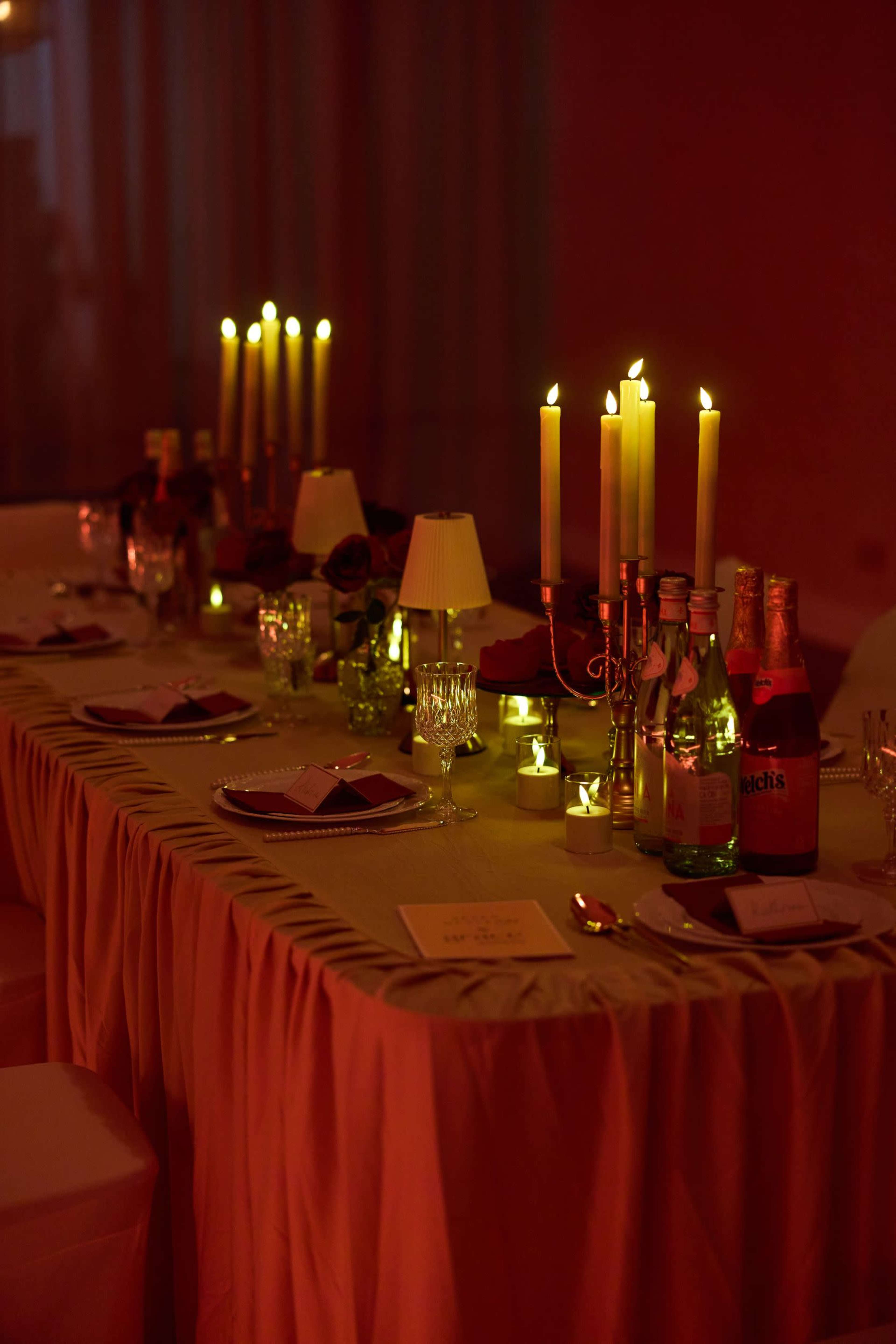 A long table is elegantly set with dim lighting, adorned with candles, bottles, and glassware for an intimate gathering.