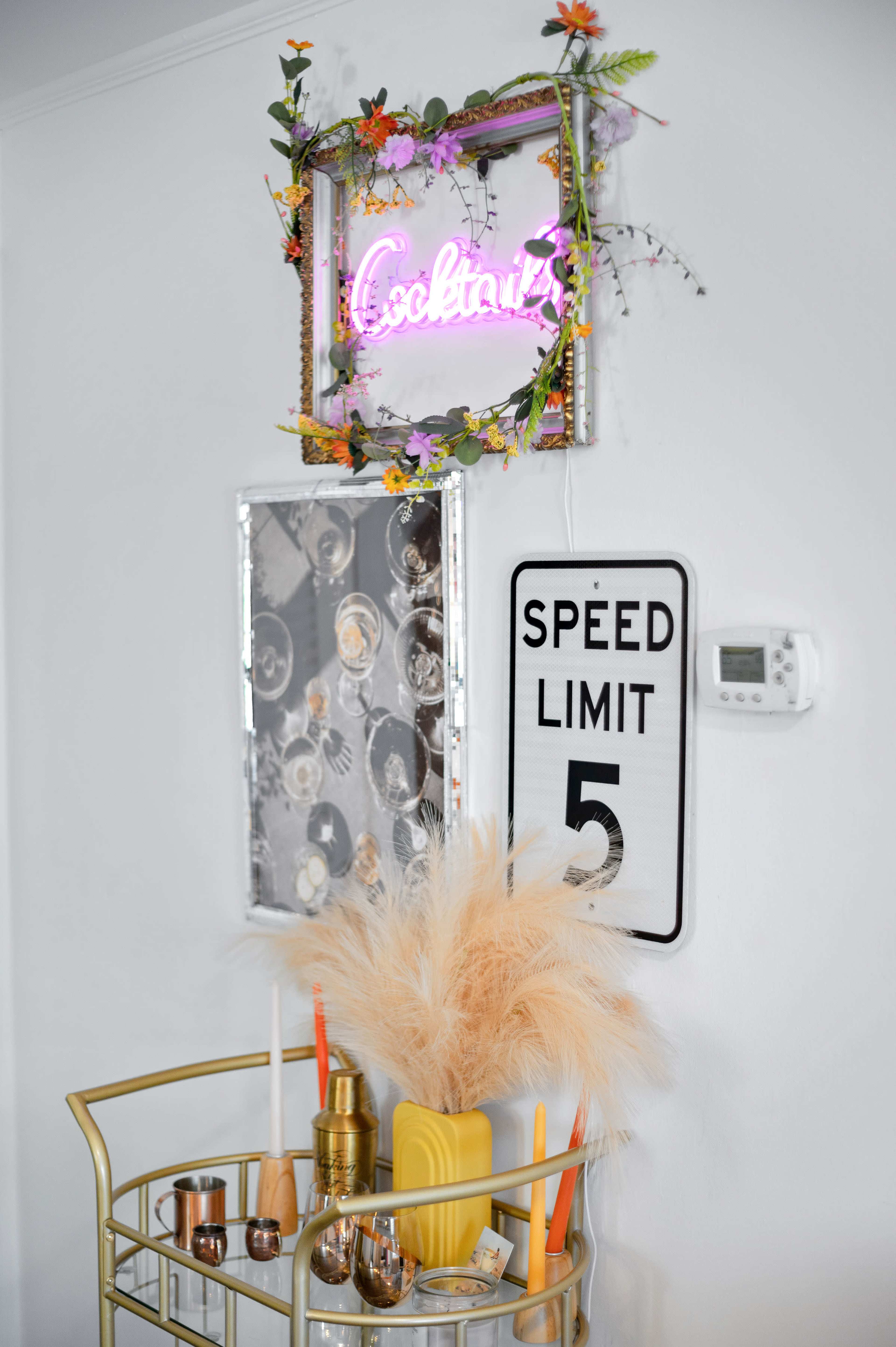 A gold bar cart displays decorative items, including candles and a yellow vase, against a wall adorned with a neon "Cocktails" sign and a speed limit sign.