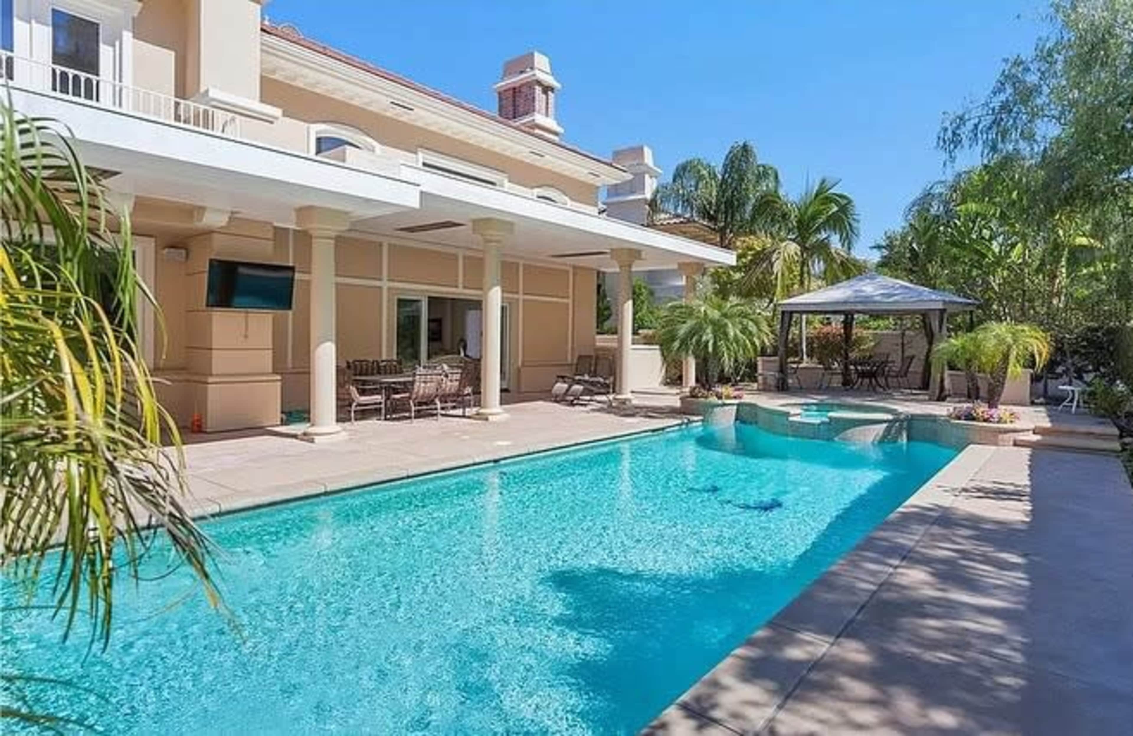 The image shows a spacious backyard with a swimming pool bordered by palm trees and a patio with outdoor seating.