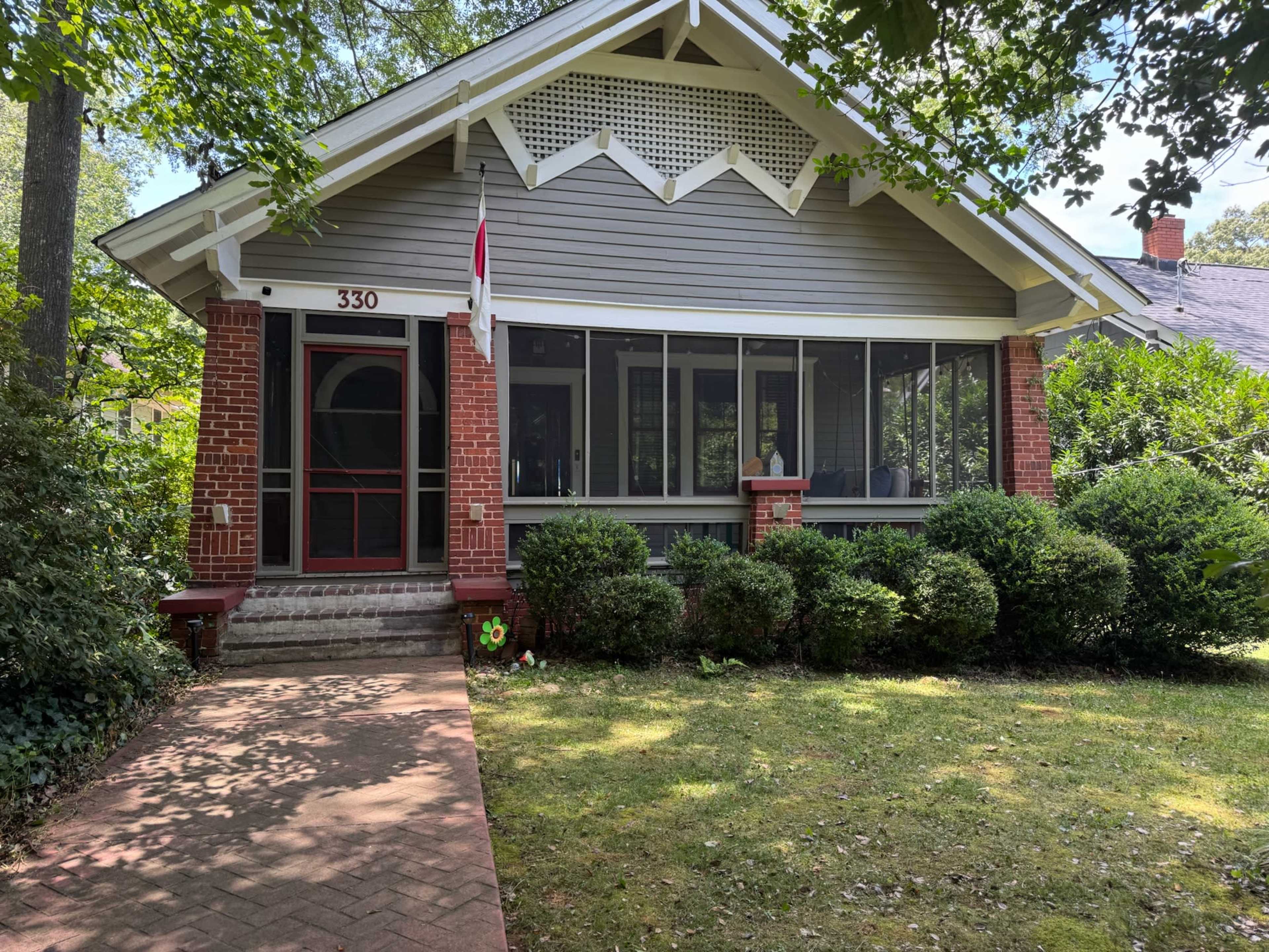 The image shows a single-story house with a screened porch, red brick details, and a well-maintained lawn surrounded by hedges.