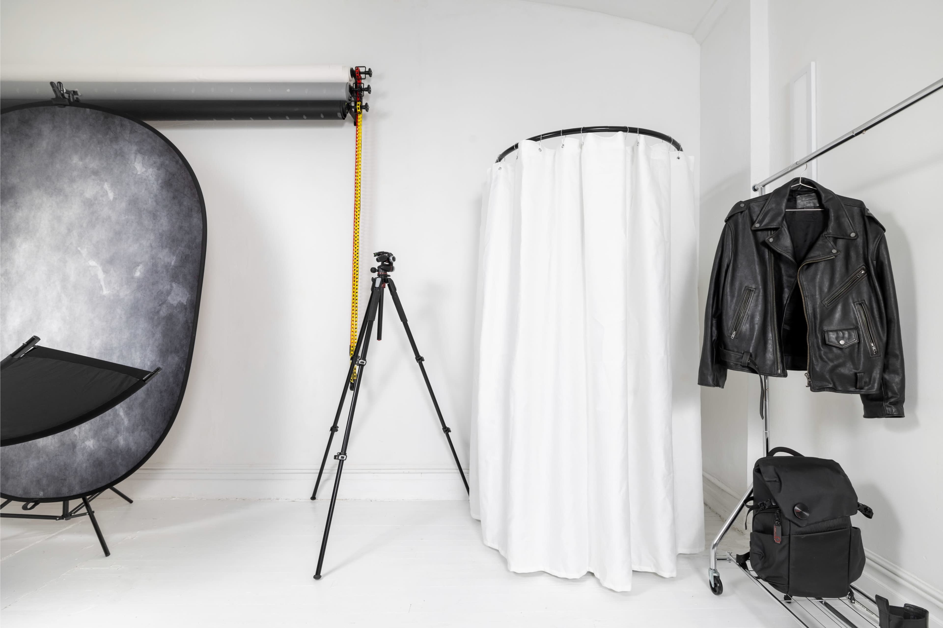 The image shows a photography studio setup featuring a backdrop, a tripod, a white curtain, a leather jacket, and a camera bag on a rack.