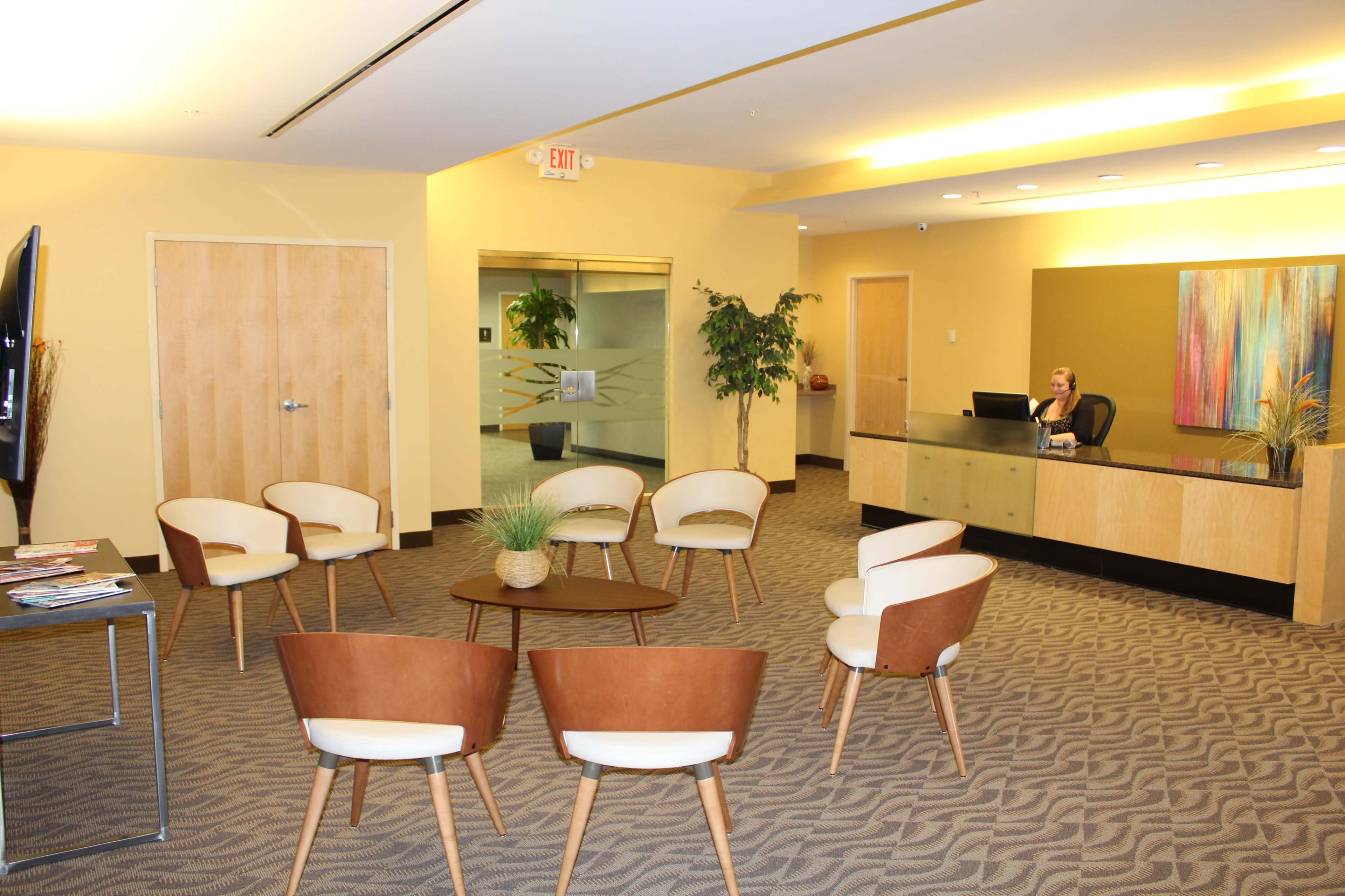 A modern reception area features a front desk with a seated receptionist, surrounded by several circular seating arrangements and a small coffee table.