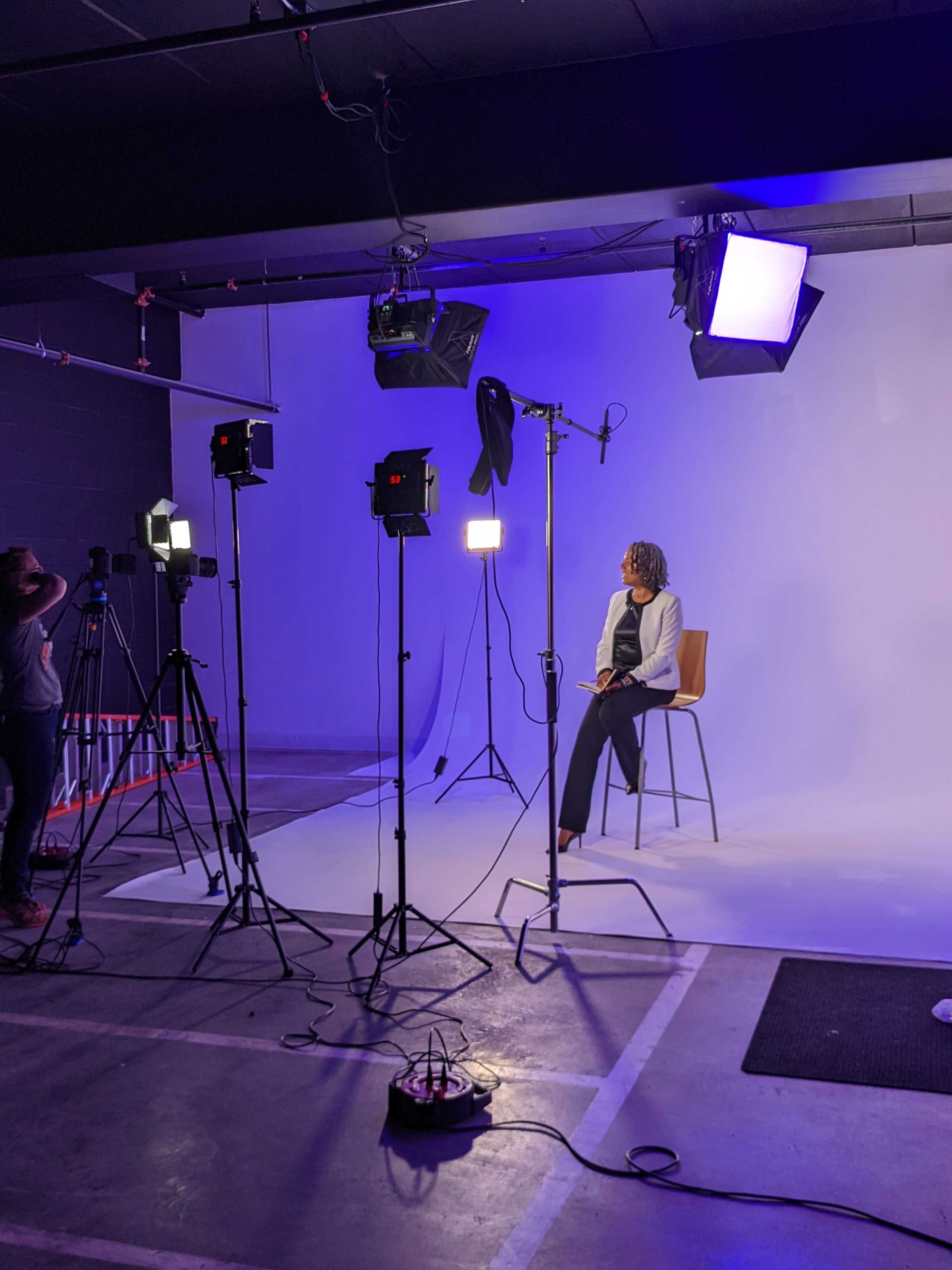 A person sits on a chair in a well-lit studio setting, surrounded by multiple lighting setups and a plain backdrop.