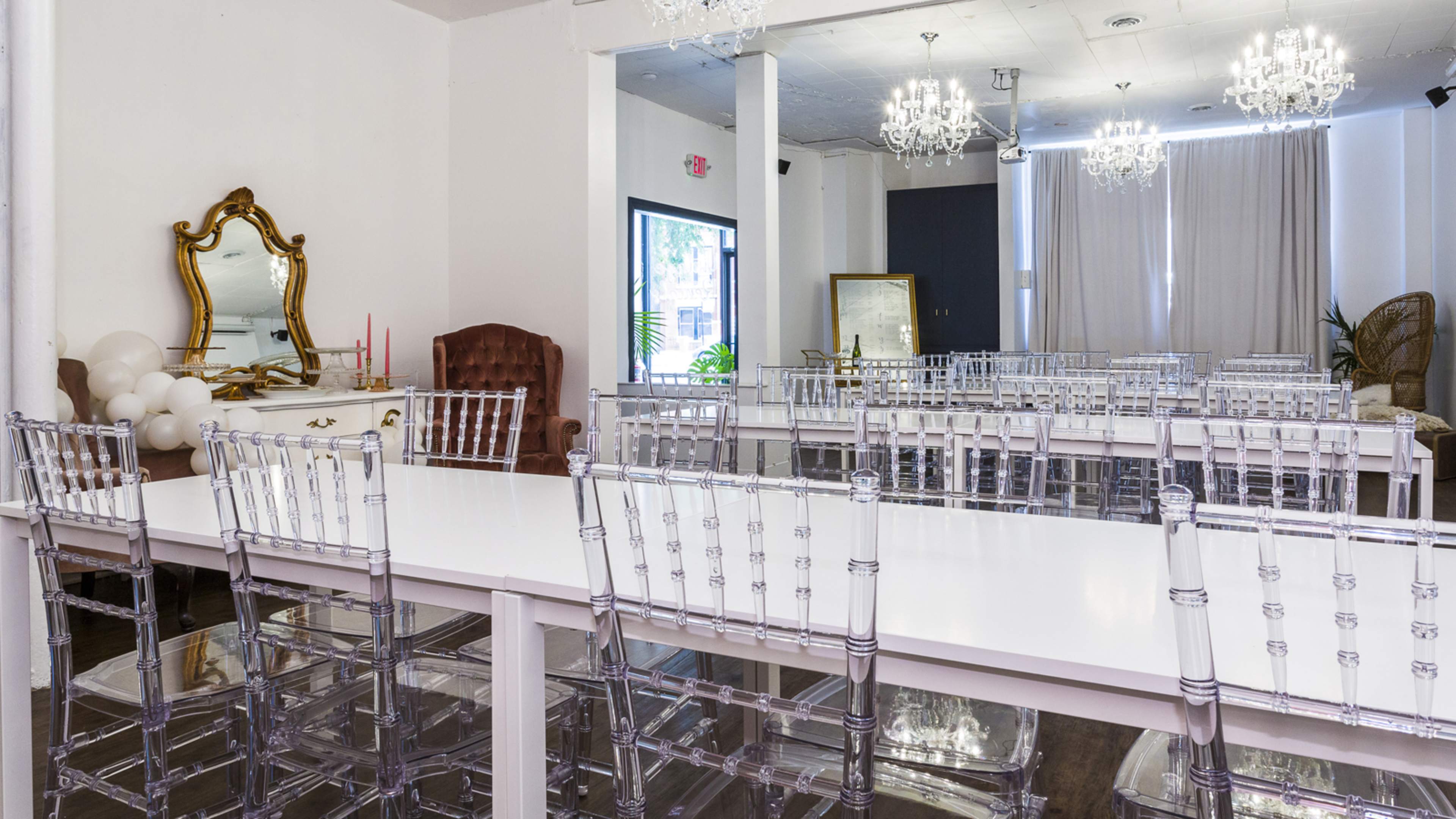 A bright event space set up with transparent chairs around long white tables, featuring decorative mirrors and chandeliers.