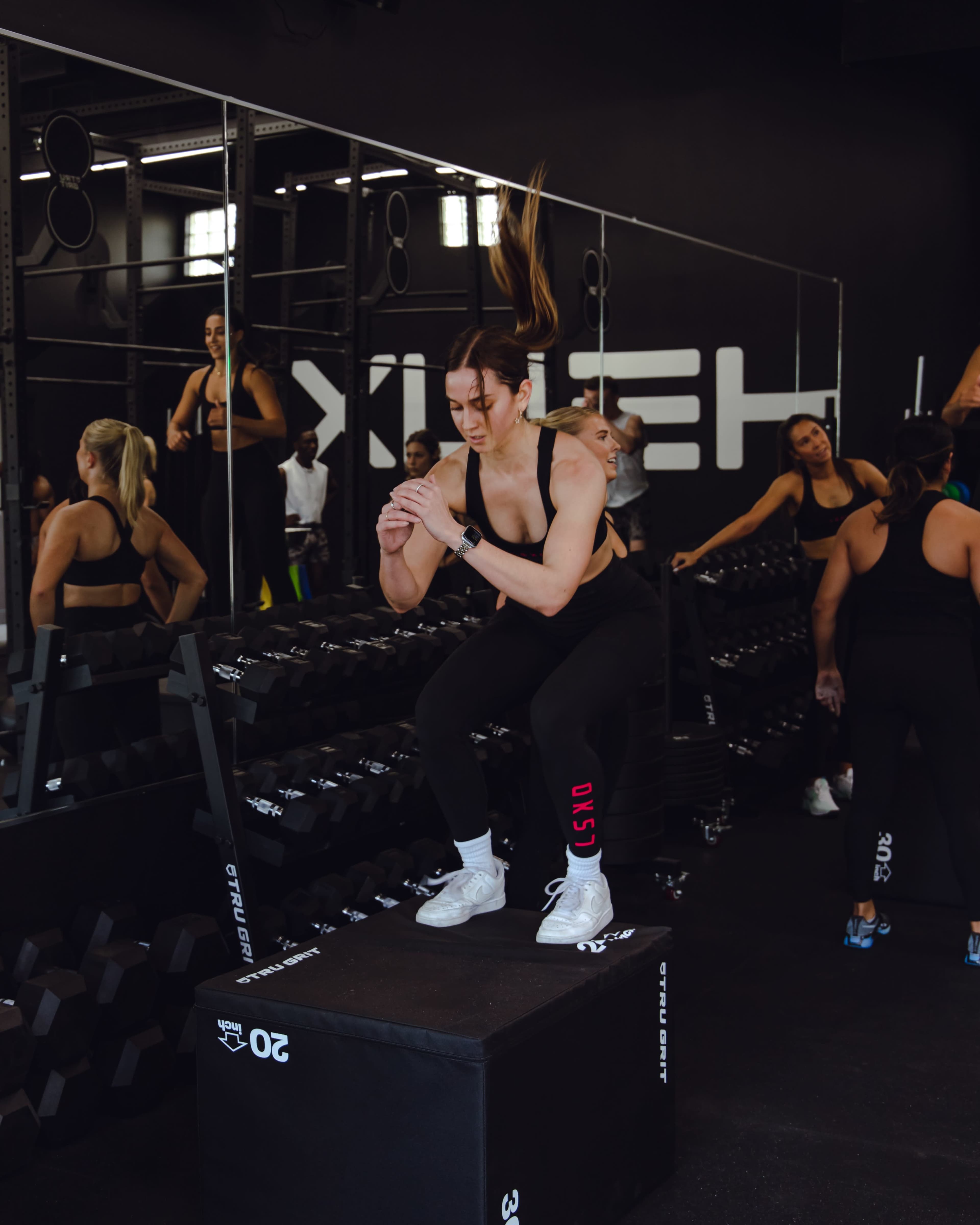 A woman performs a box jump on a raised platform in a gym filled with weights and other individuals exercising.