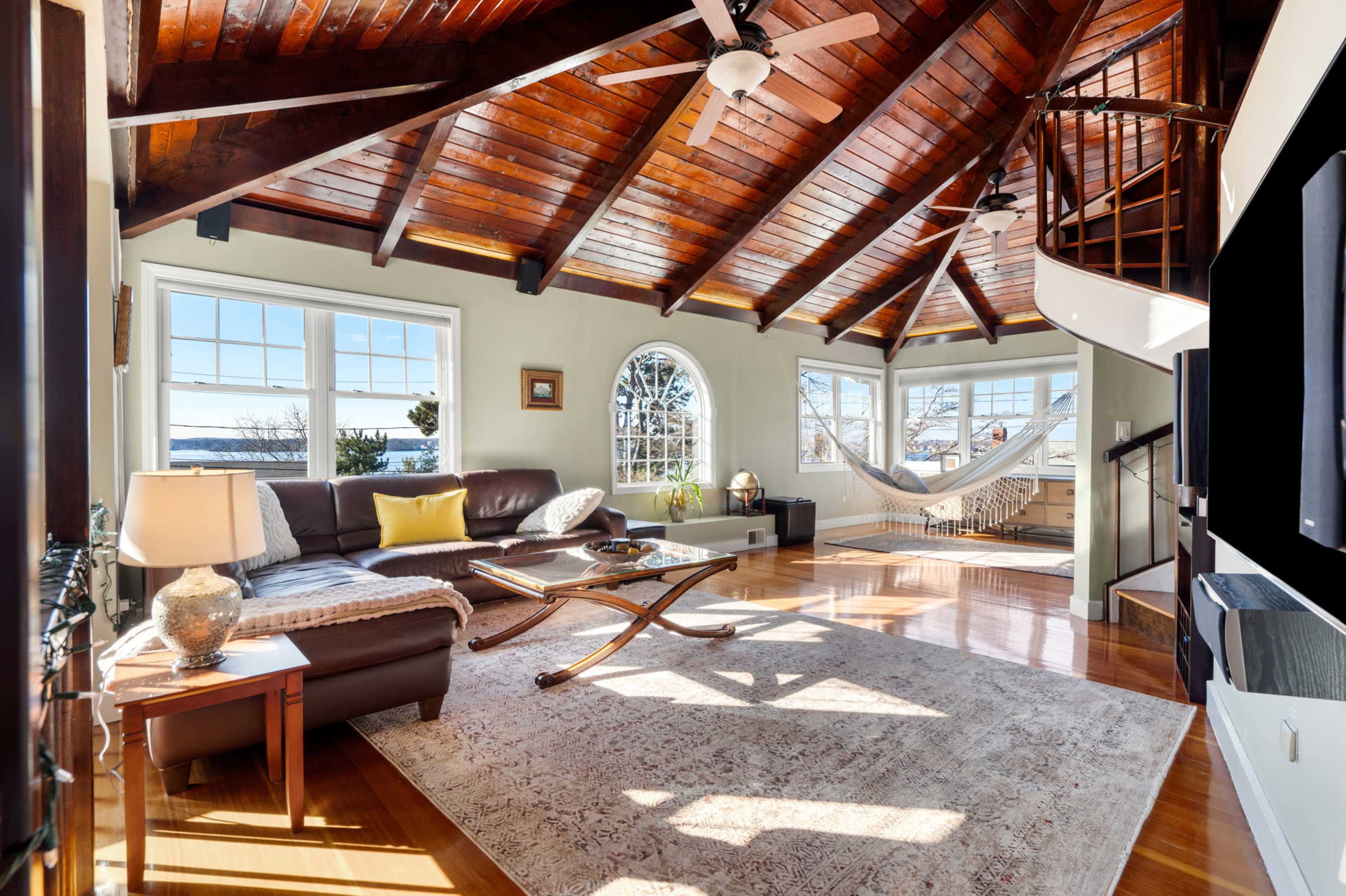 A spacious living room with a high wooden ceiling, large windows, a comfortable couch, and a spiral staircase leading to an upper level.