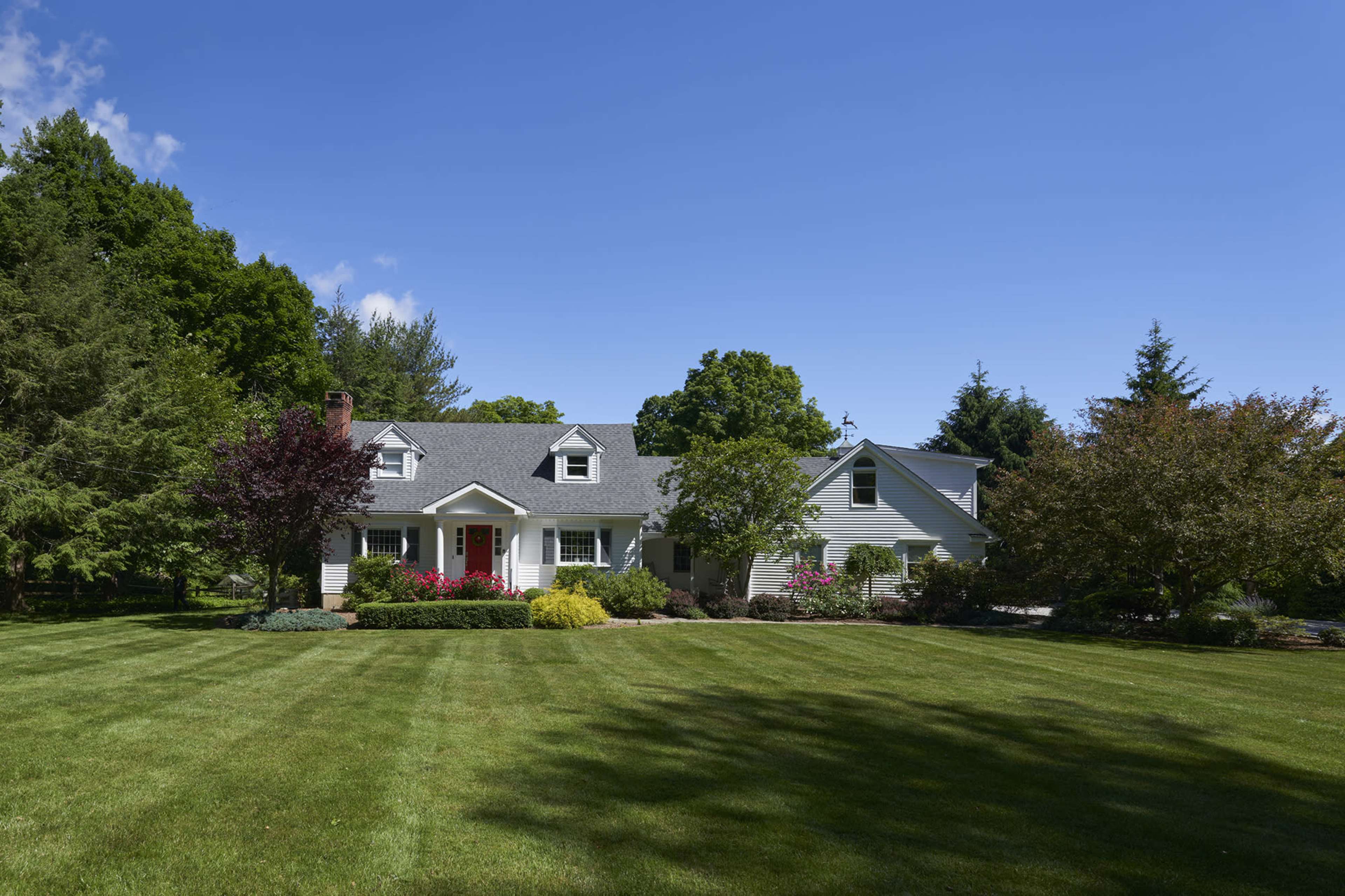 A white house with a gray roof is surrounded by a well-maintained lawn and trees under a clear blue sky.