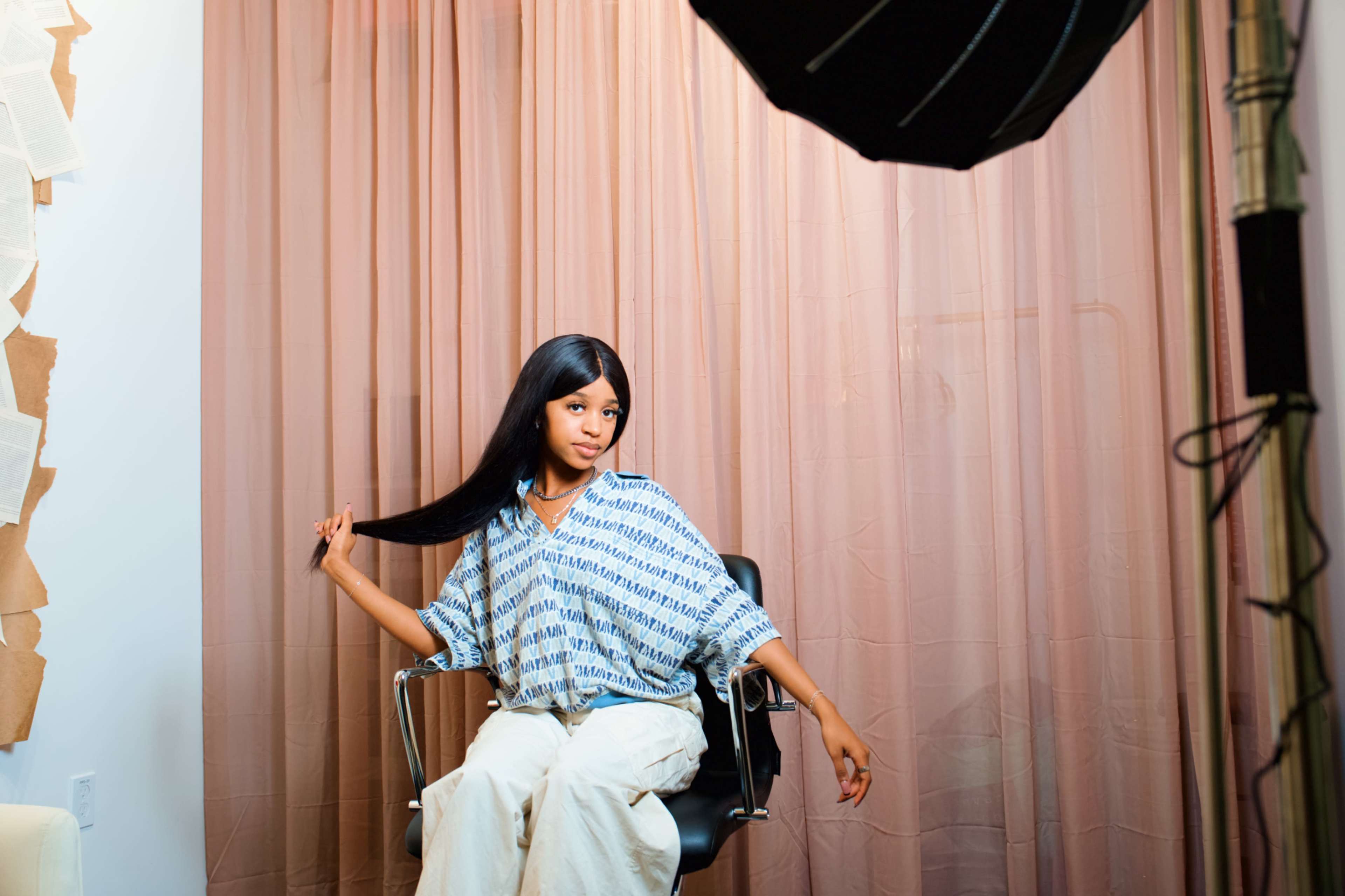 A person sits in a black chair in a studio setting with a soft fabric backdrop and a lighting setup.