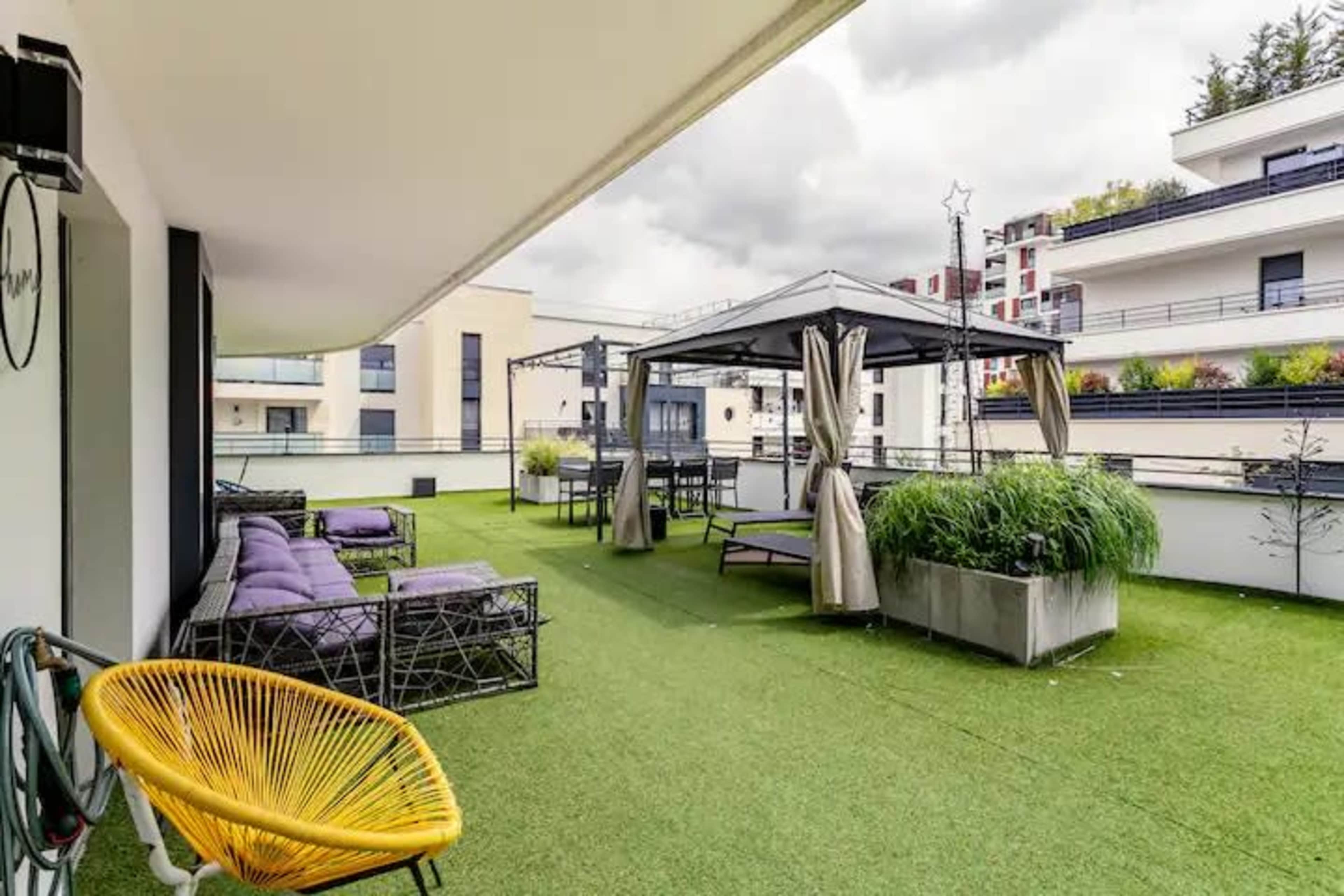 A modern rooftop terrace featuring seating areas, planters, and a gazebo, all set against a backdrop of buildings and cloudy skies.