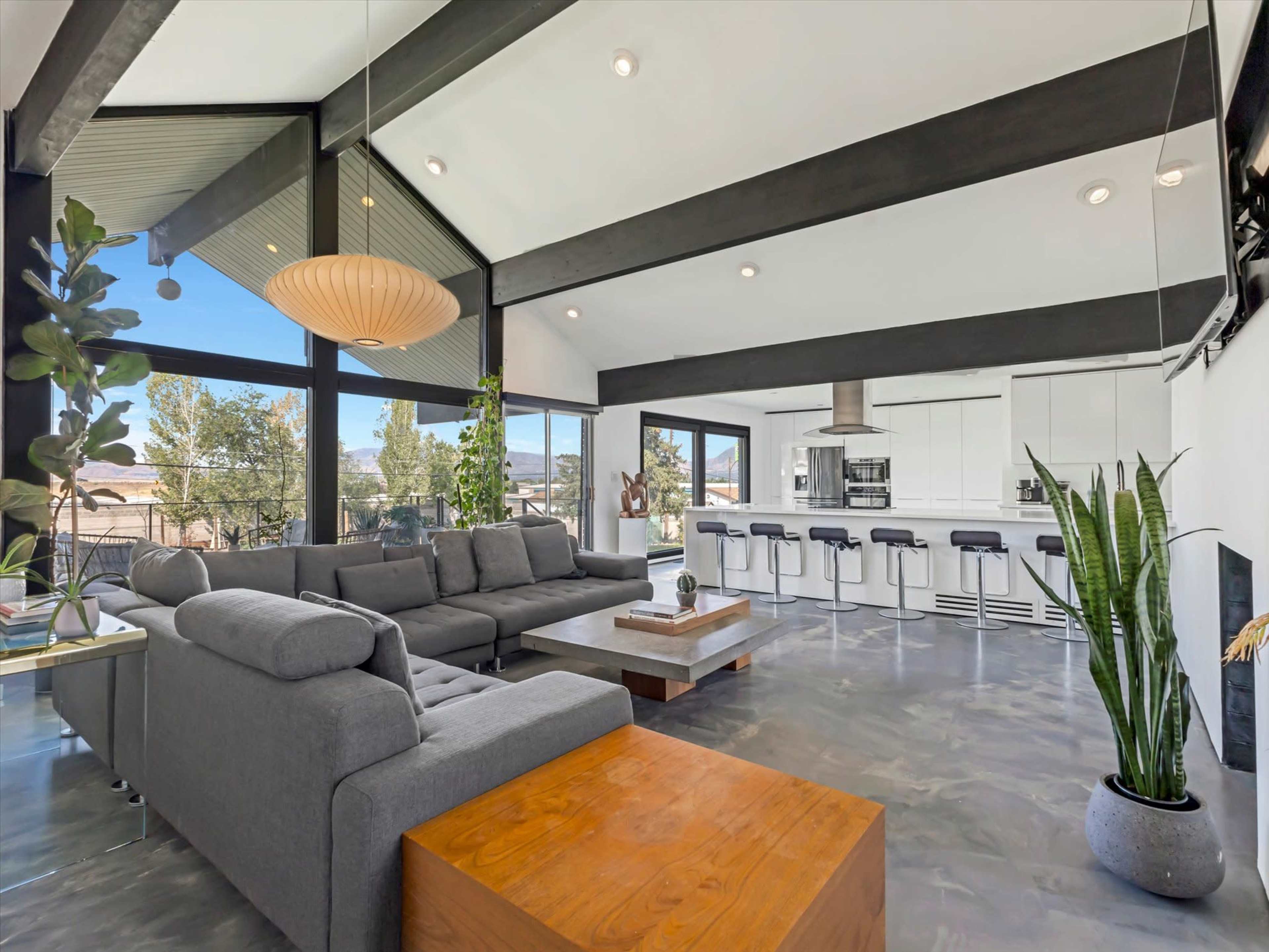 The image shows a modern interior living space with a gray sectional sofa, a wooden coffee table, and a kitchen area featuring sleek cabinetry and appliances, framed by large windows that provide natural light and a view of greenery outside.