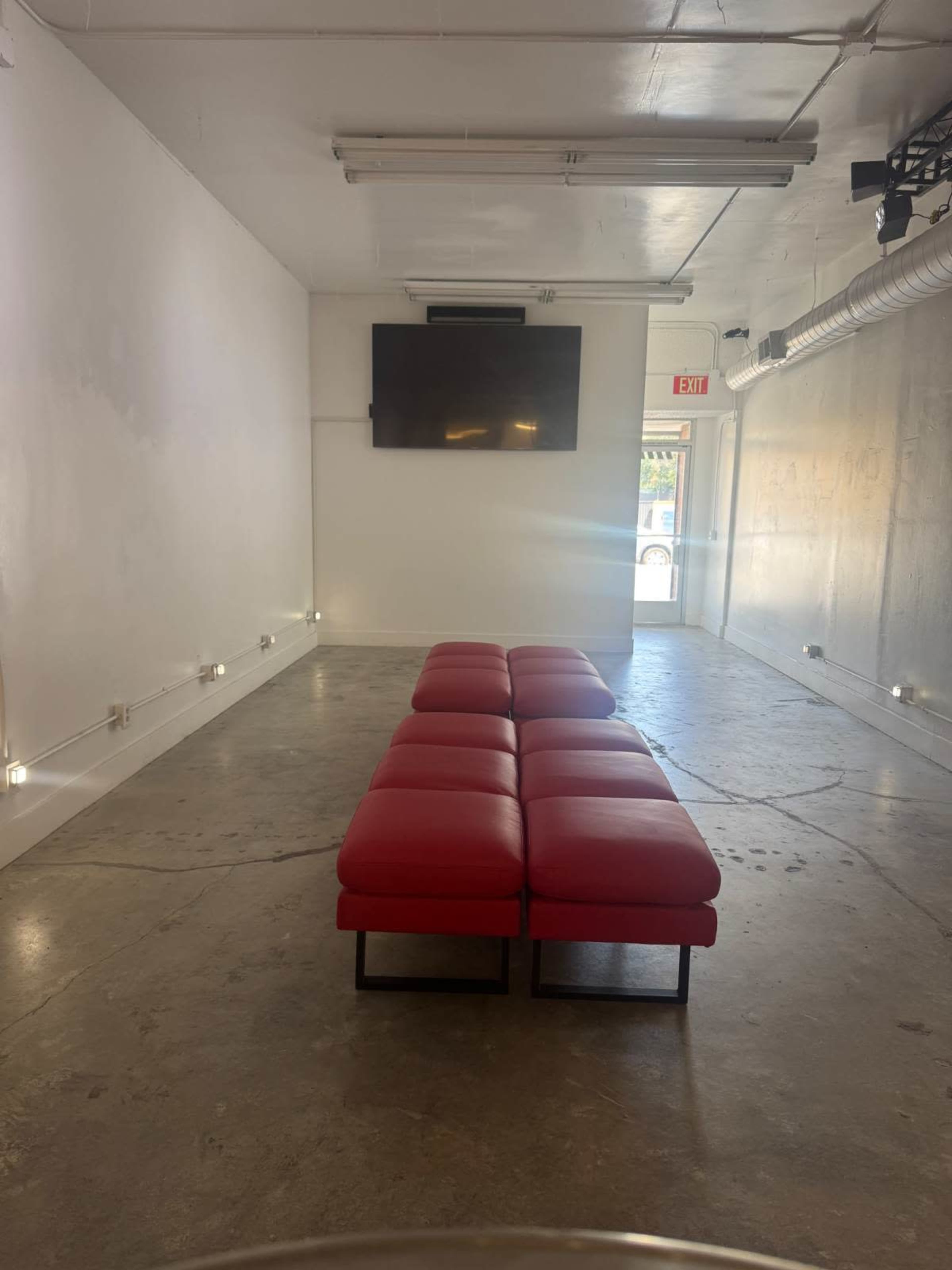 The image shows a minimalist interior space featuring a series of red benches arranged in a line, with a large wall-mounted television at one end.