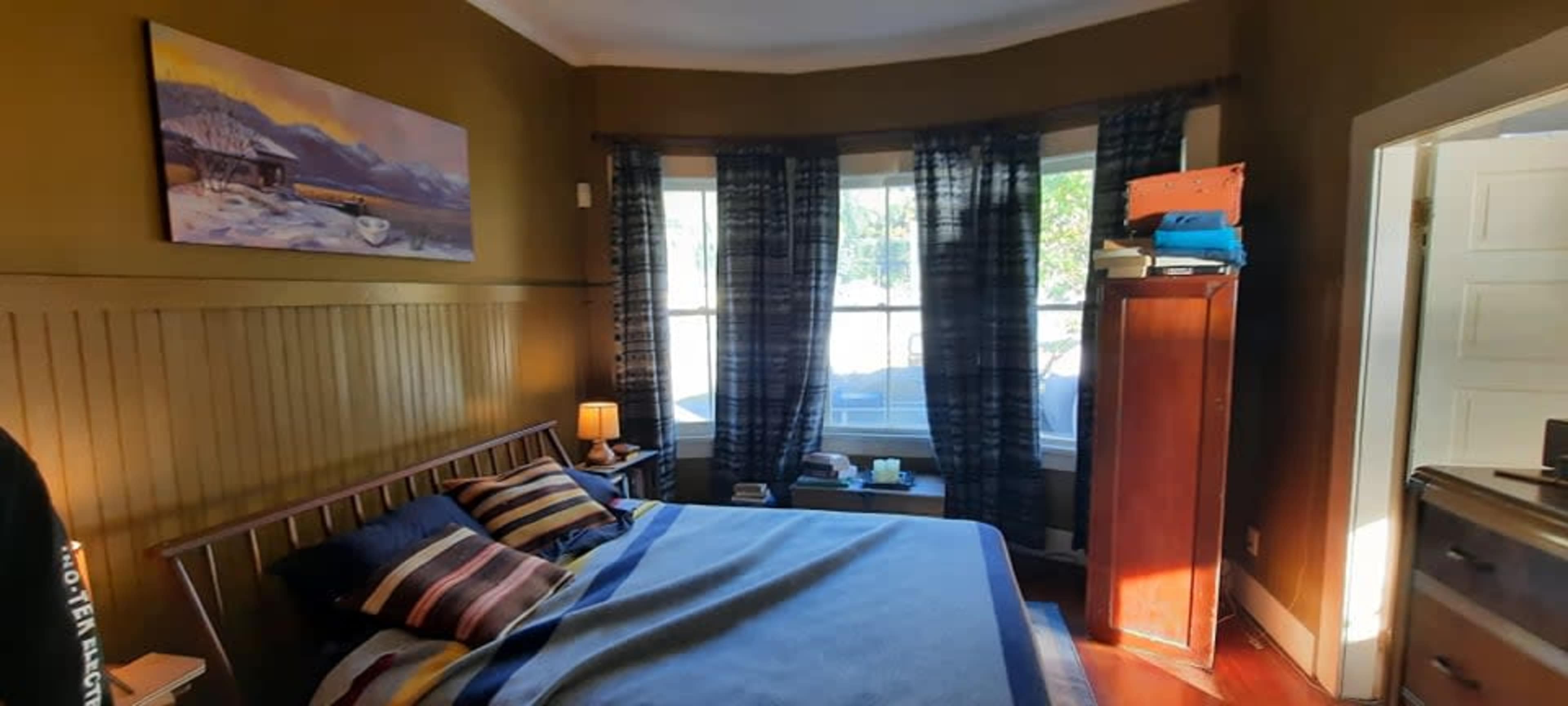 The image shows a cozy bedroom with a bed covered in striped blankets, a wooden nightstand, and large windows flanked by dark curtains.