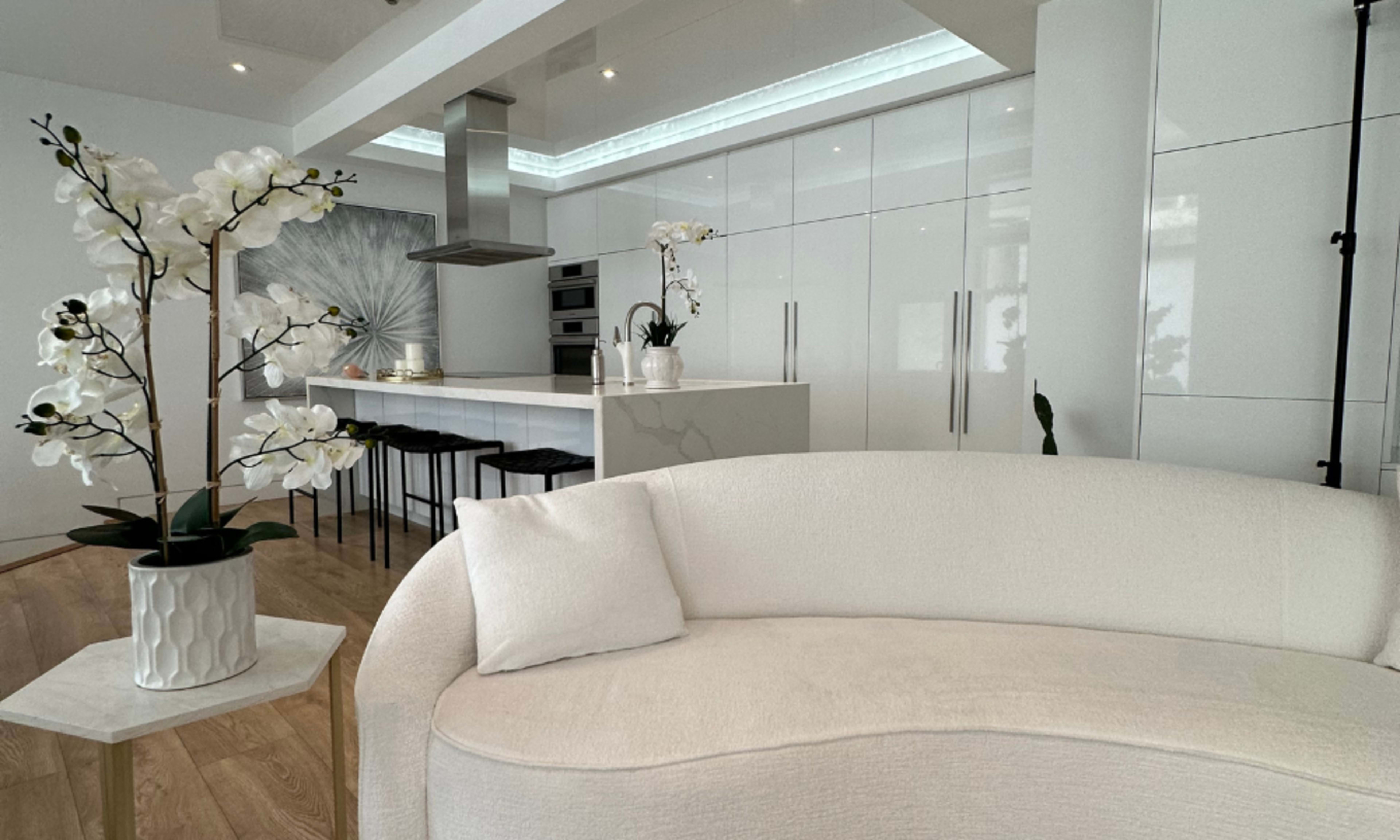A modern kitchen space features white cabinetry, a central island with bar stools, and decorative orchids in a minimalist living area.
