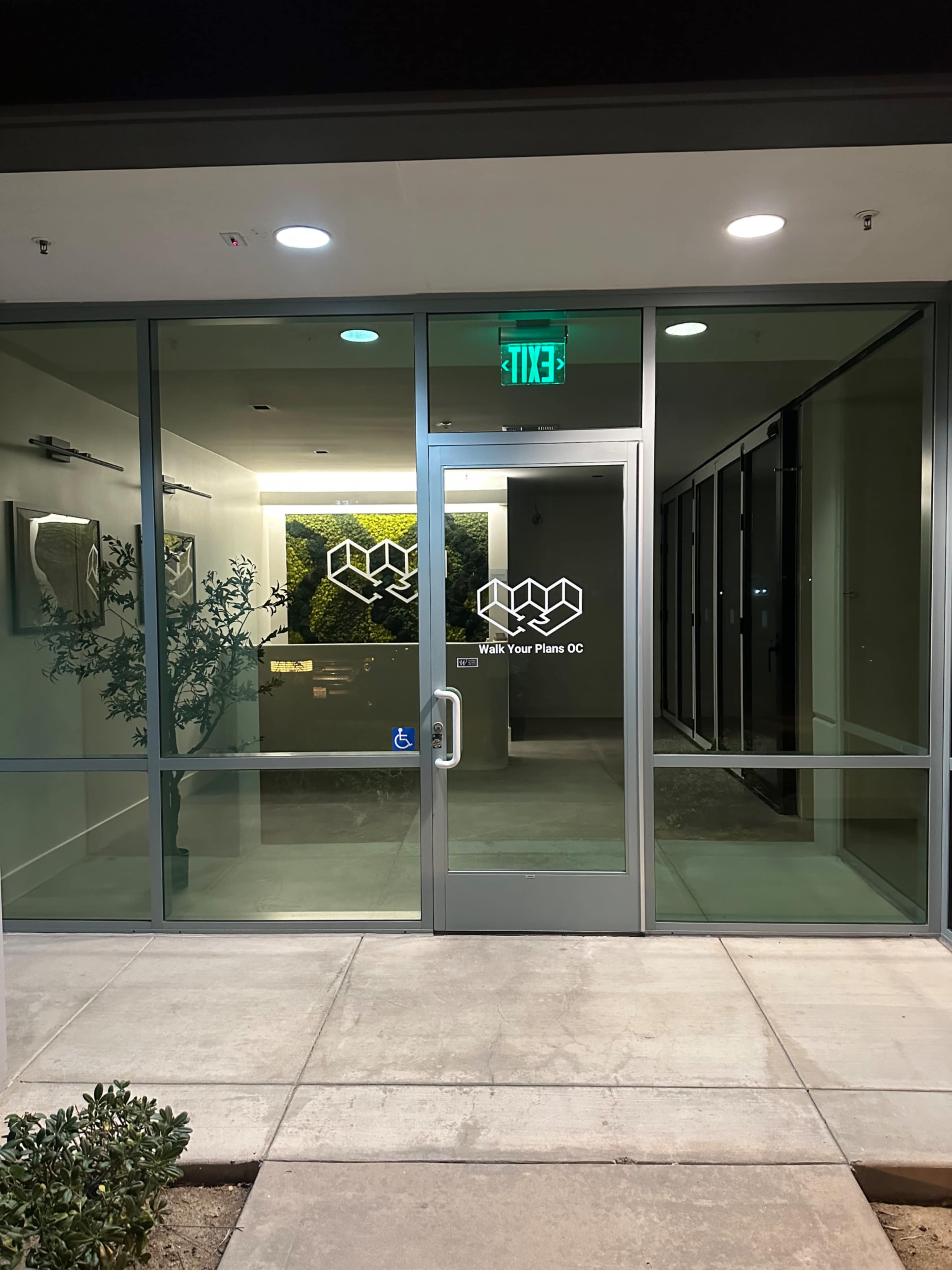 The entrance of a modern glass storefront features a double door with a logo and a sign that reads "Walk Your Plans OC."