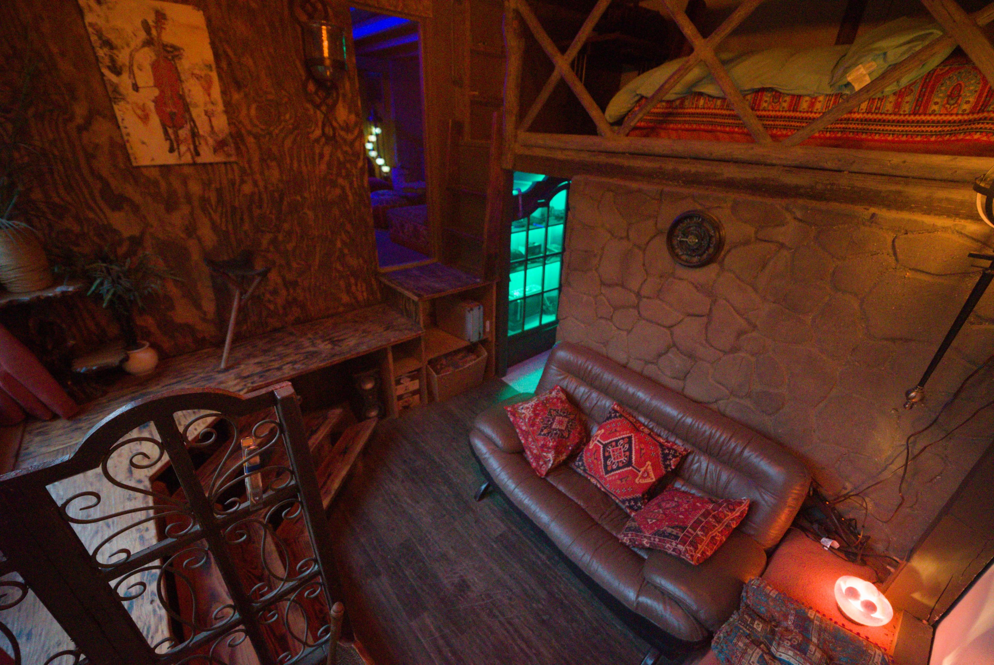 The image shows a cozy interior of a small space featuring a lofted bed, a sofa with decorative pillows, and textured walls illuminated by soft lighting.