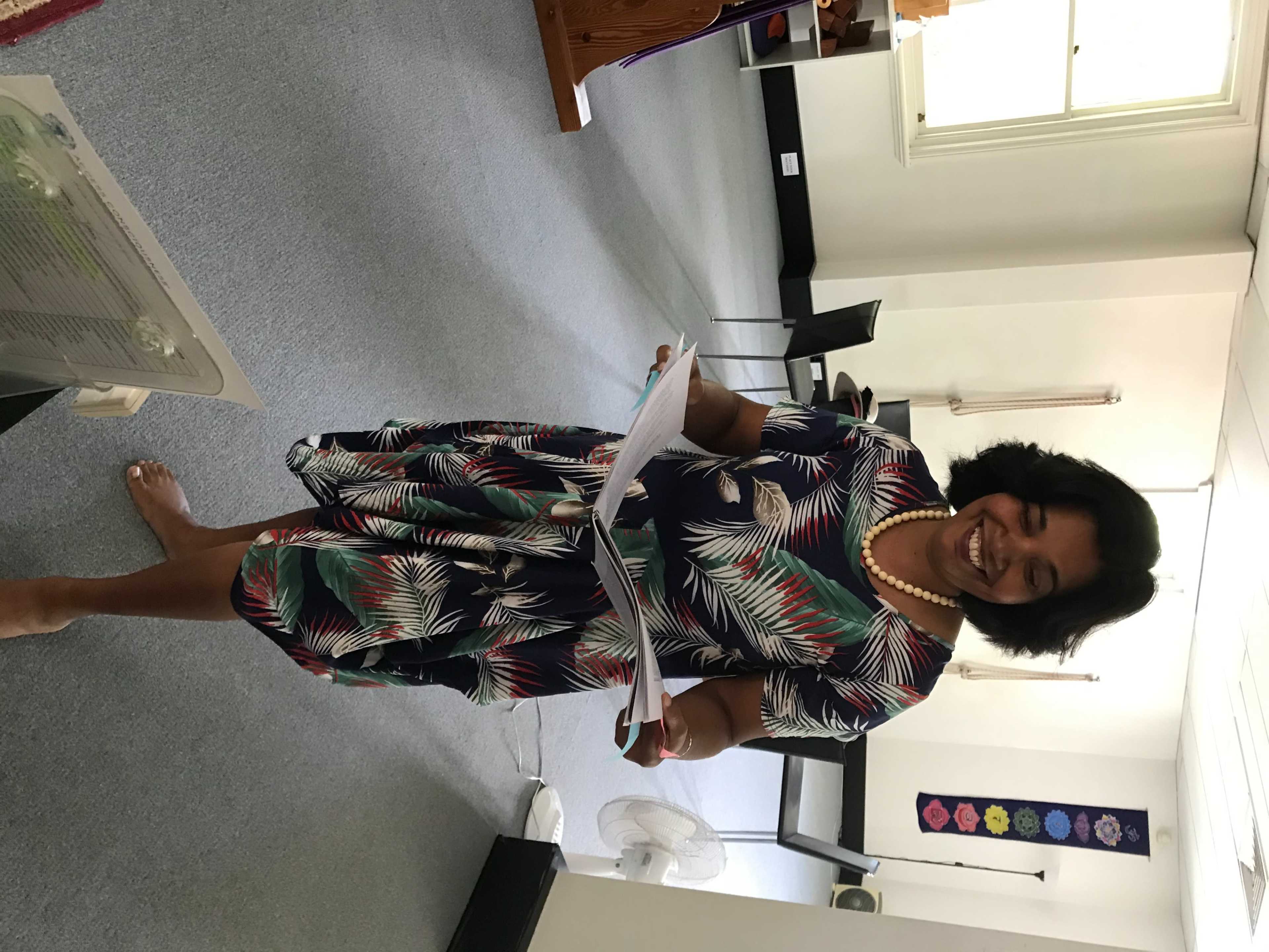 A woman in a floral dress stands barefoot in a room holding papers and smiling.