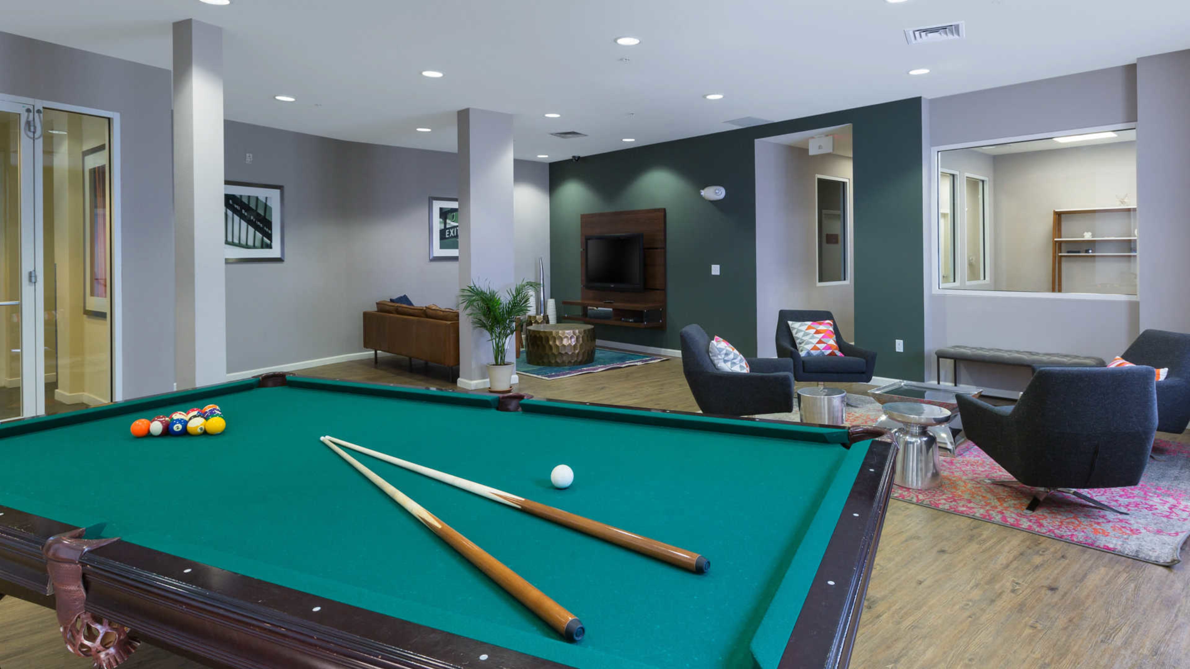 A game room features a pool table in the foreground, with a comfortable seating area and a television in the background.