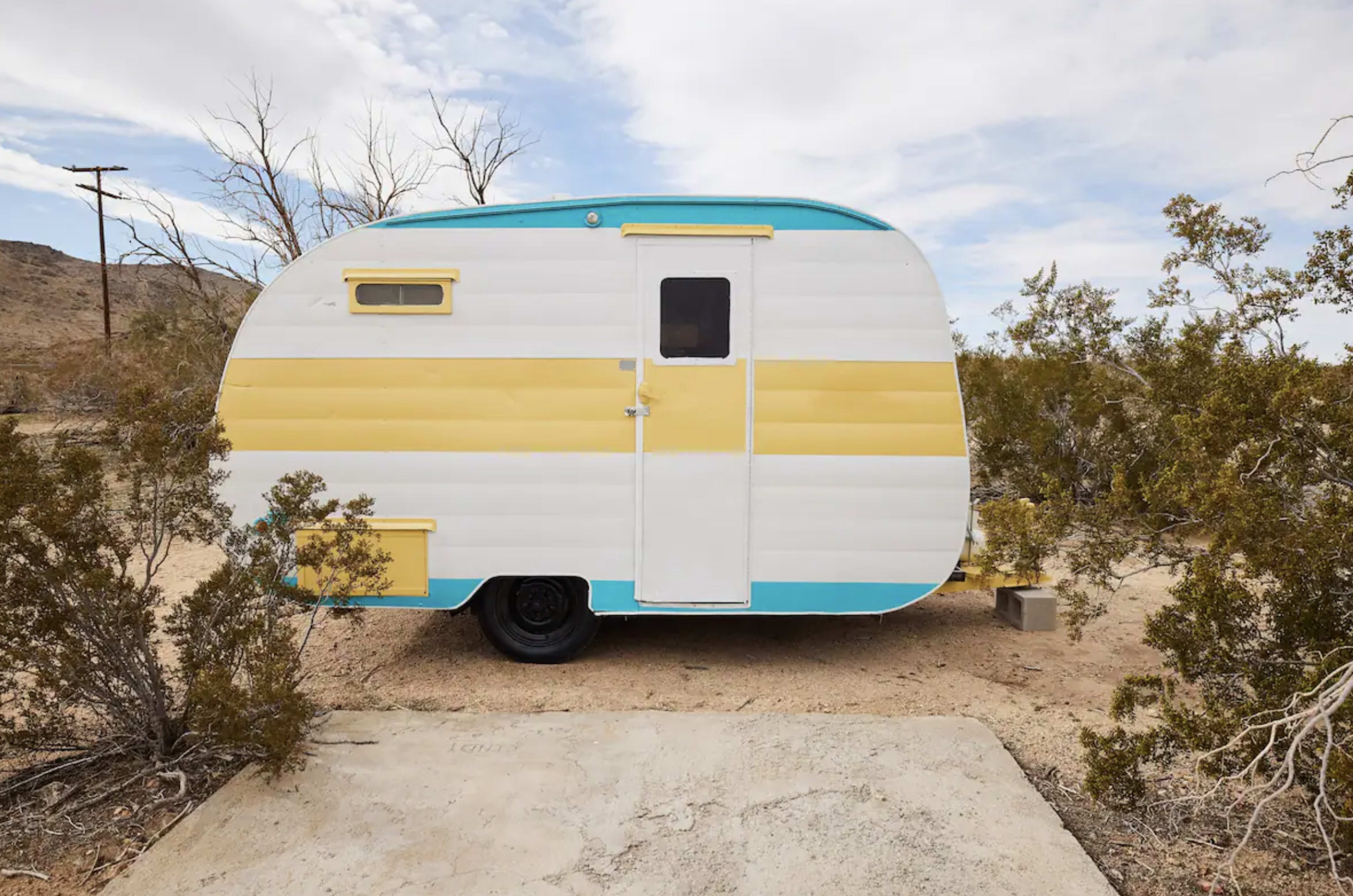 A vintage travel trailer with a yellow and blue exterior is parked on a dusty patch of ground surrounded by sparse vegetation in a desert landscape.