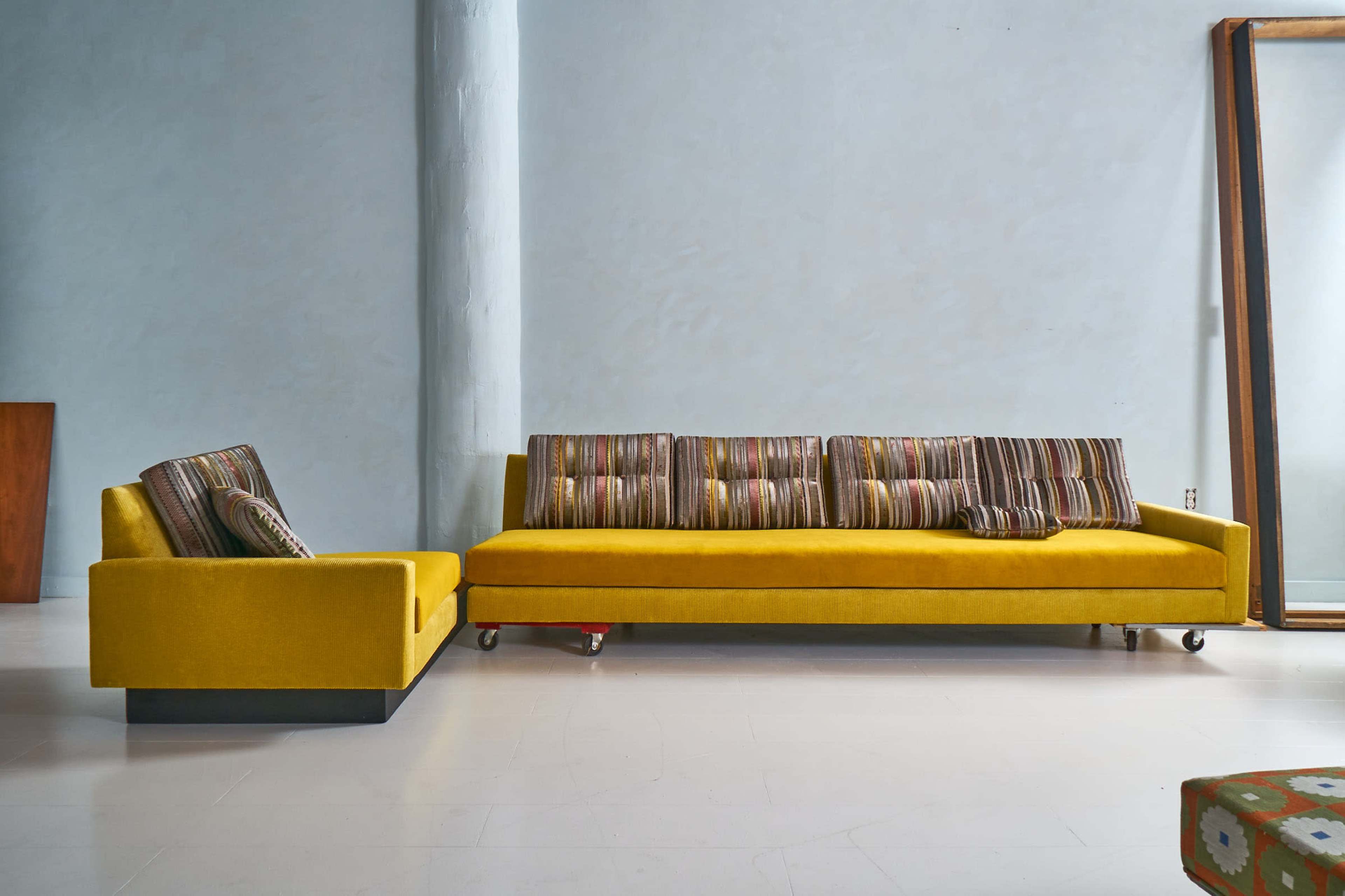 A yellow sectional sofa with striped cushions is placed against a light blue wall in a minimalist room.