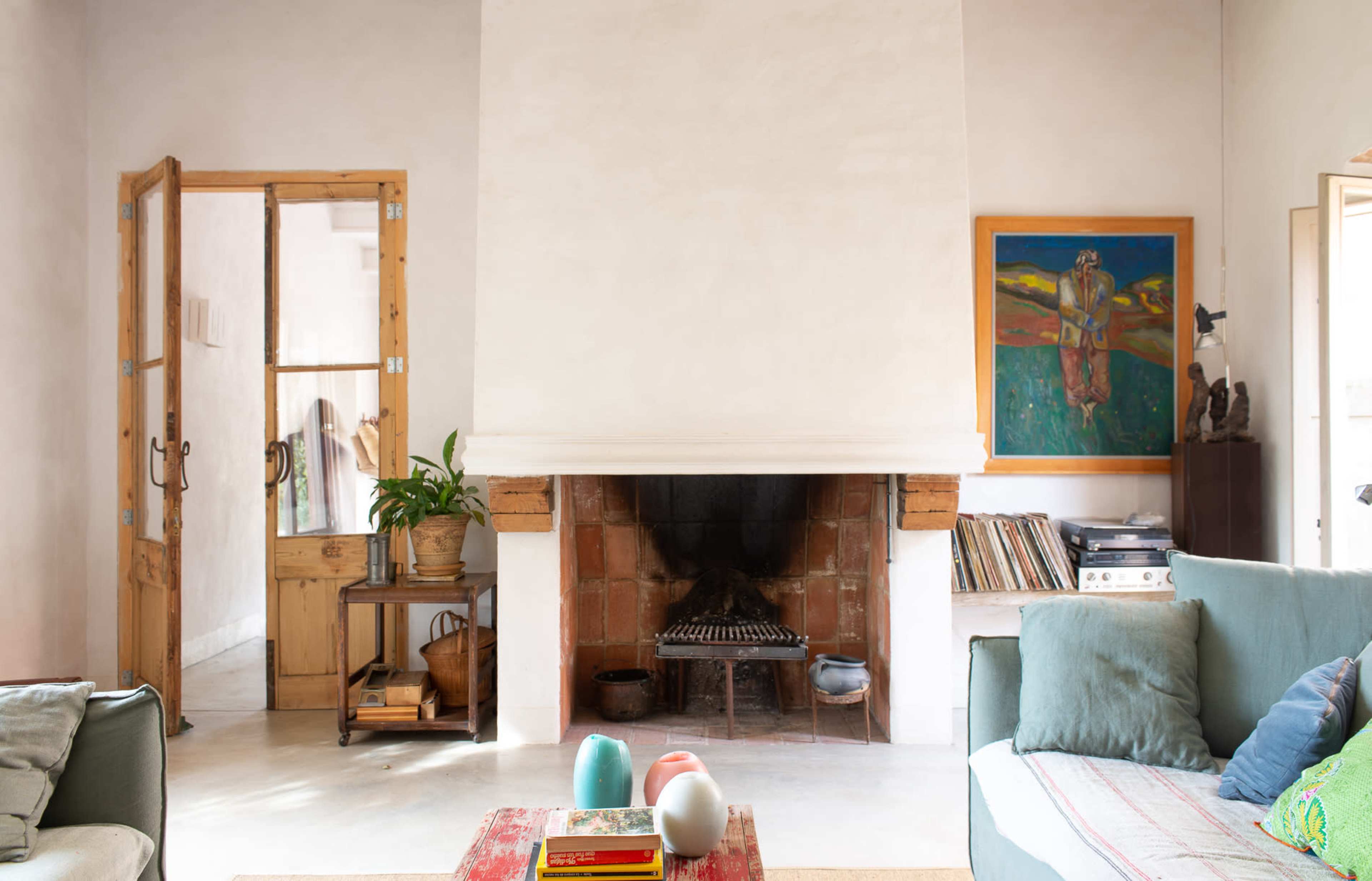 A cozy living room features a central fireplace, a bookshelf, and a door leading to another room, with a green sofa and decorative items on a low table.