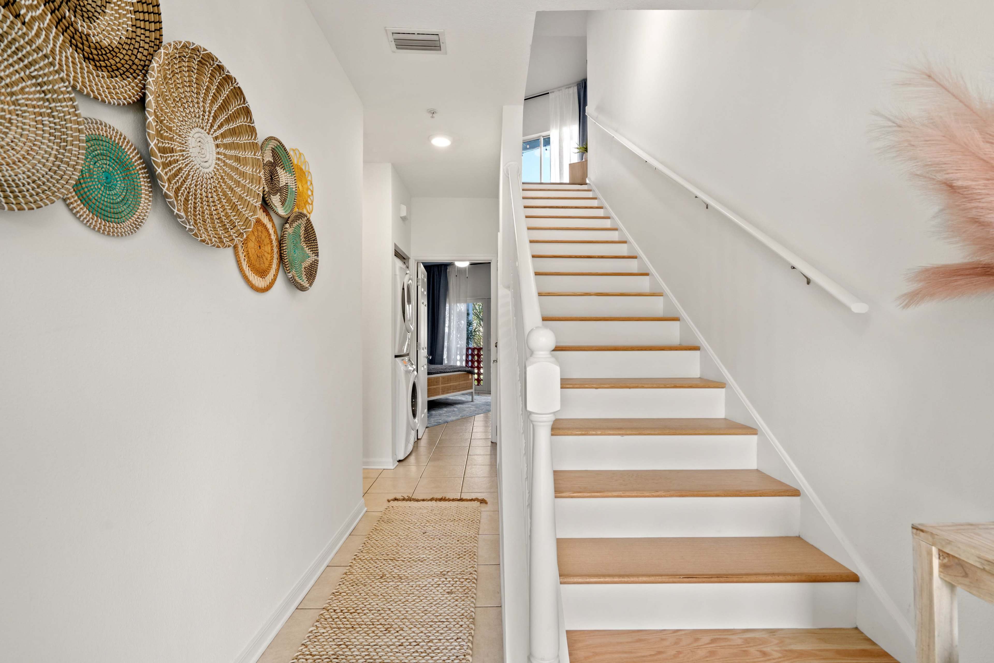 A brightly lit hallway features a staircase leading to the upper level, with decorative woven baskets on the wall and a beige runner mat on the floor.