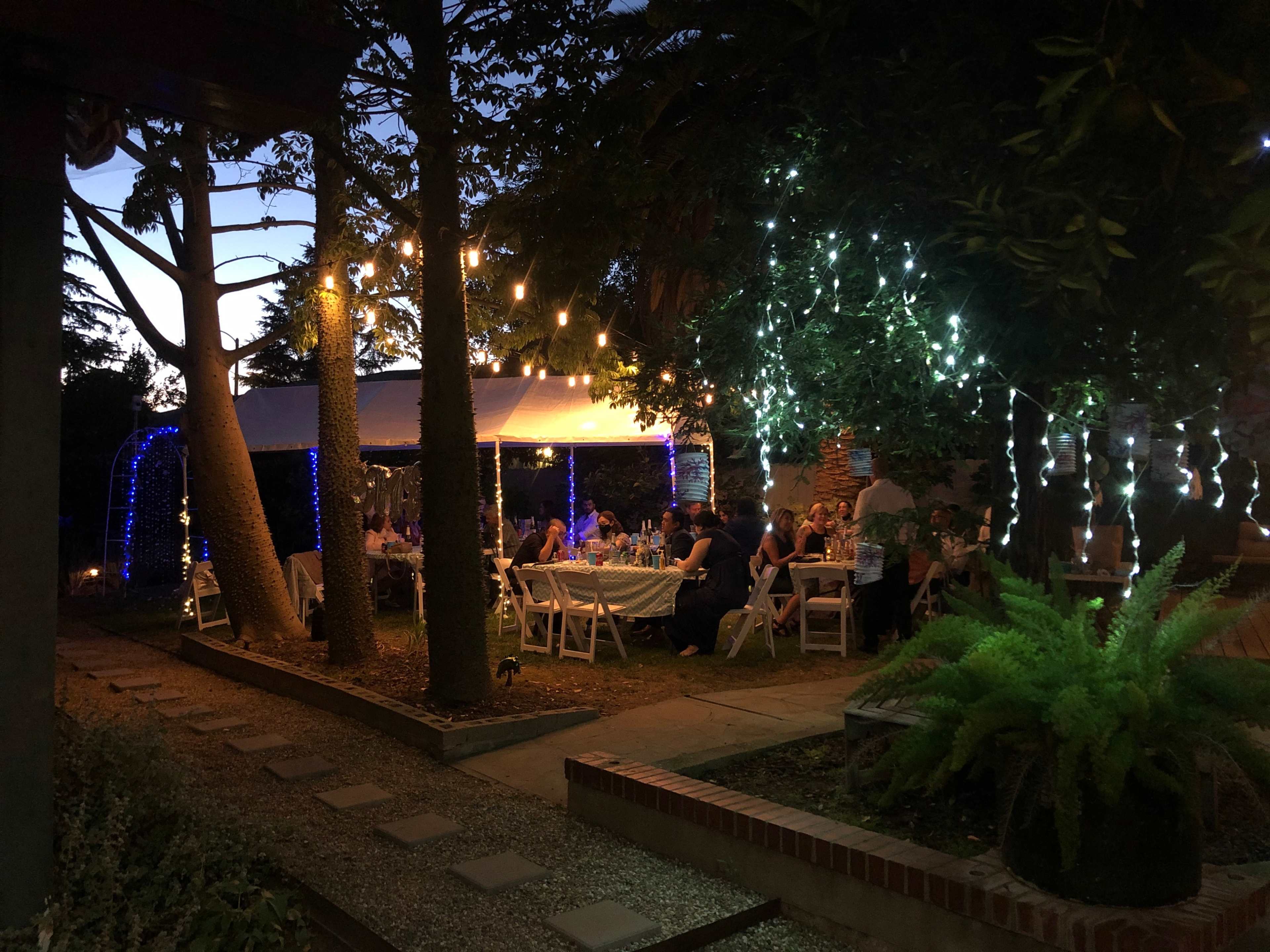The scene shows a well-lit outdoor gathering under string lights with tables set for dining among trees and greenery at dusk.