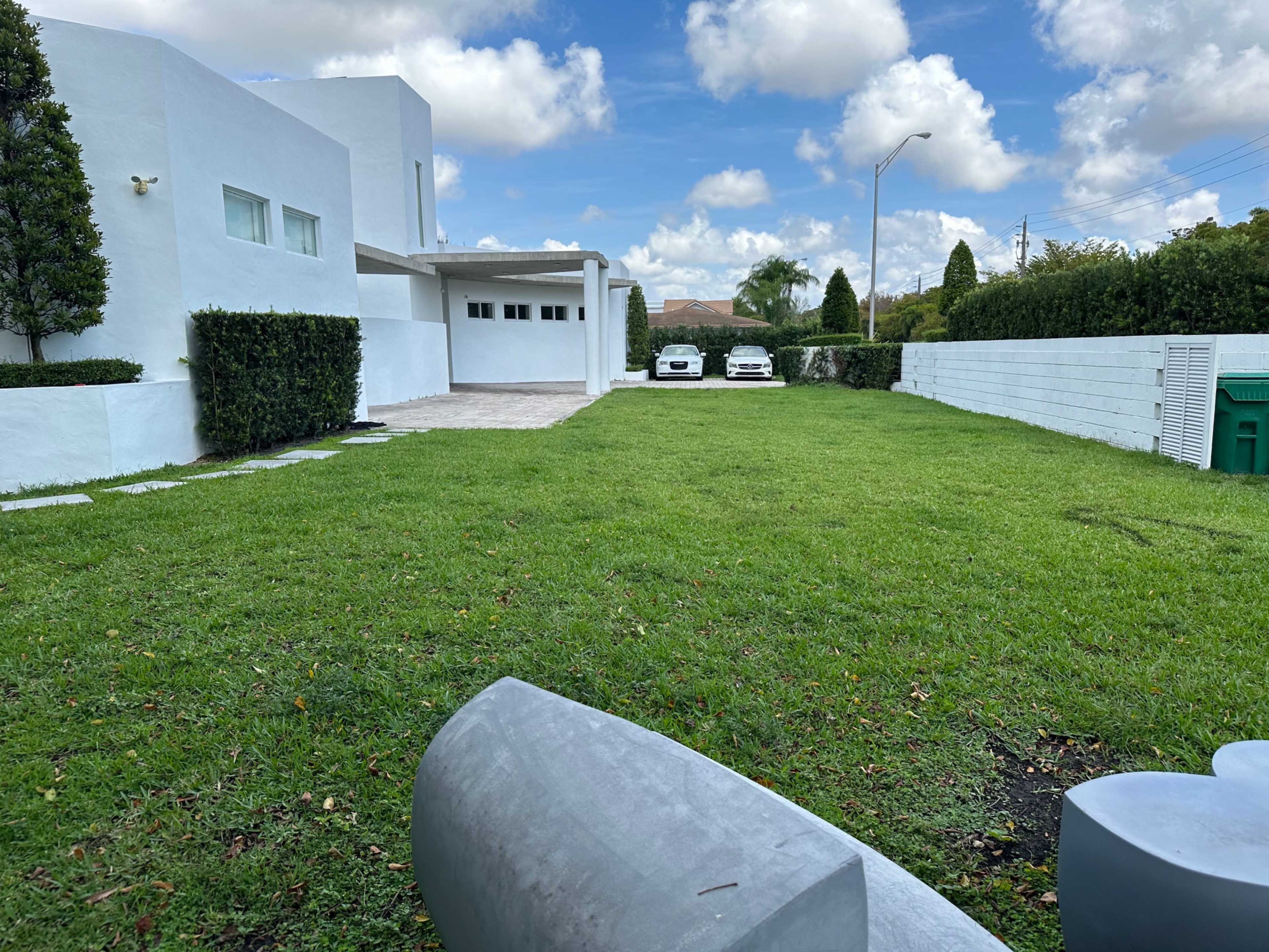 A green lawn stretches between a white modern house and two parked cars in a driveway, with a low wall and shrubs lining the property.