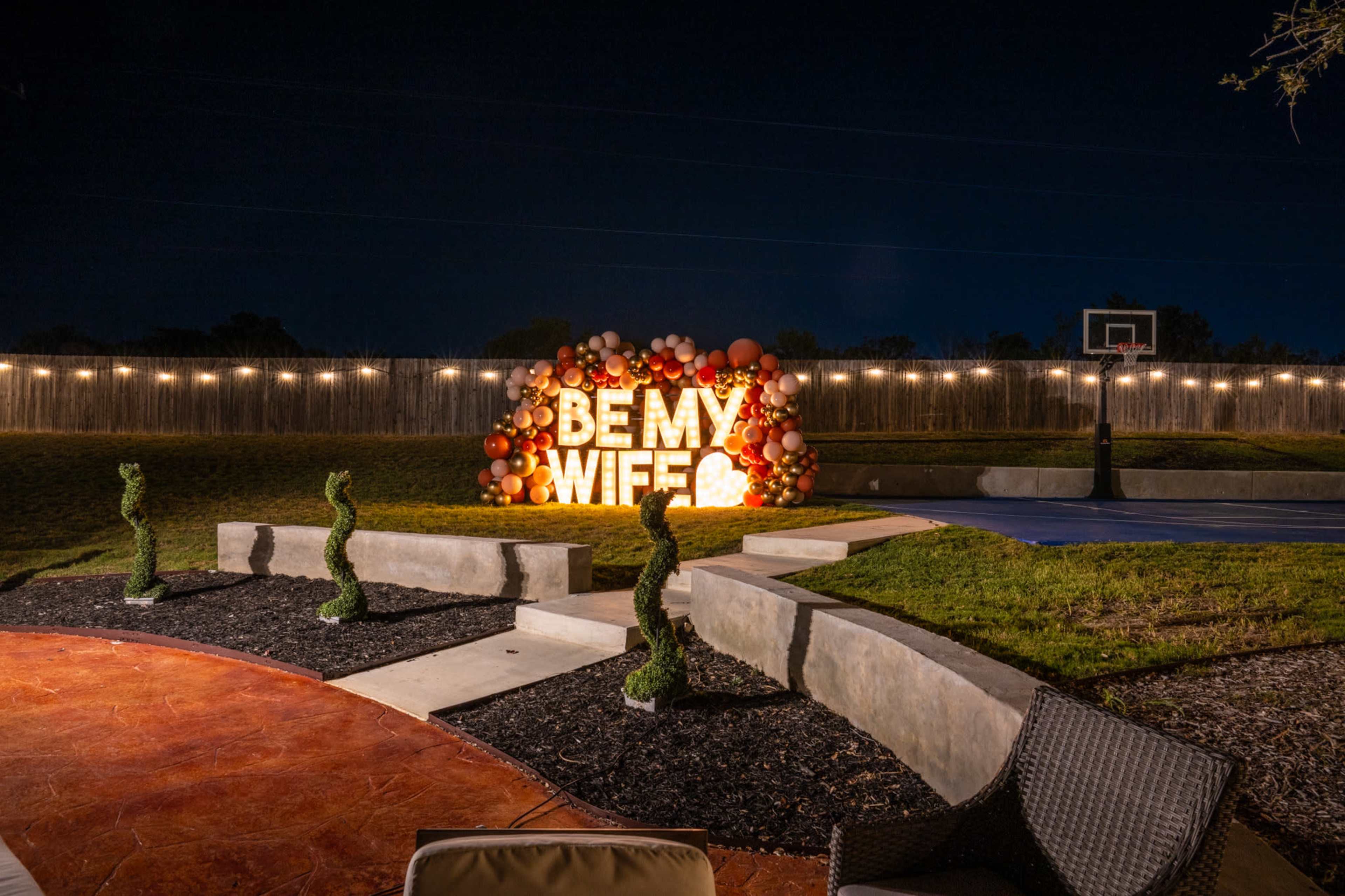 A brightly lit display reading "BE MY WIFE" is surrounded by a colorful balloon arrangement in a backyard at night.