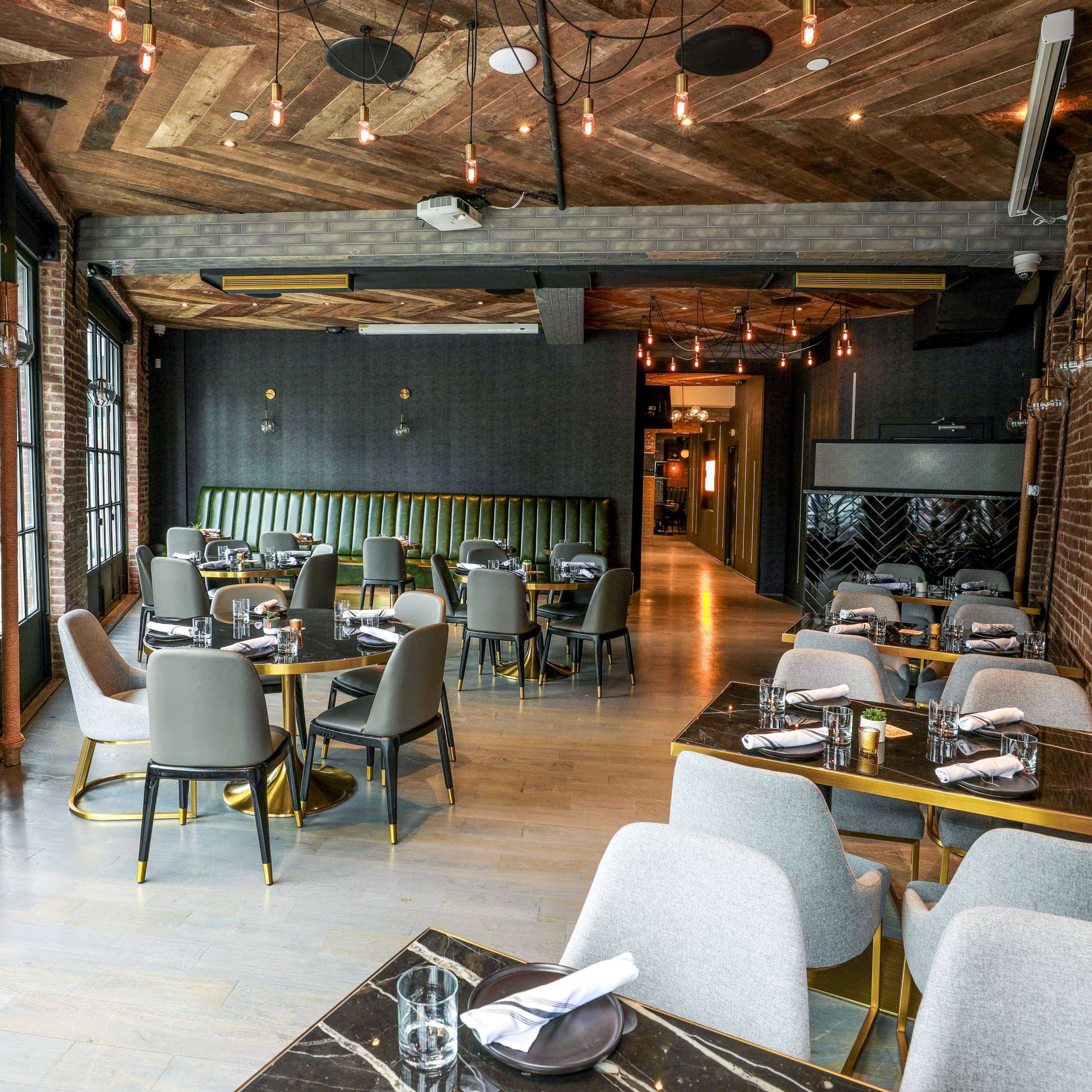 The image shows a modern restaurant interior featuring round tables set with utensils, grey upholstered chairs, and a distinctive wooden ceiling with hanging lights.