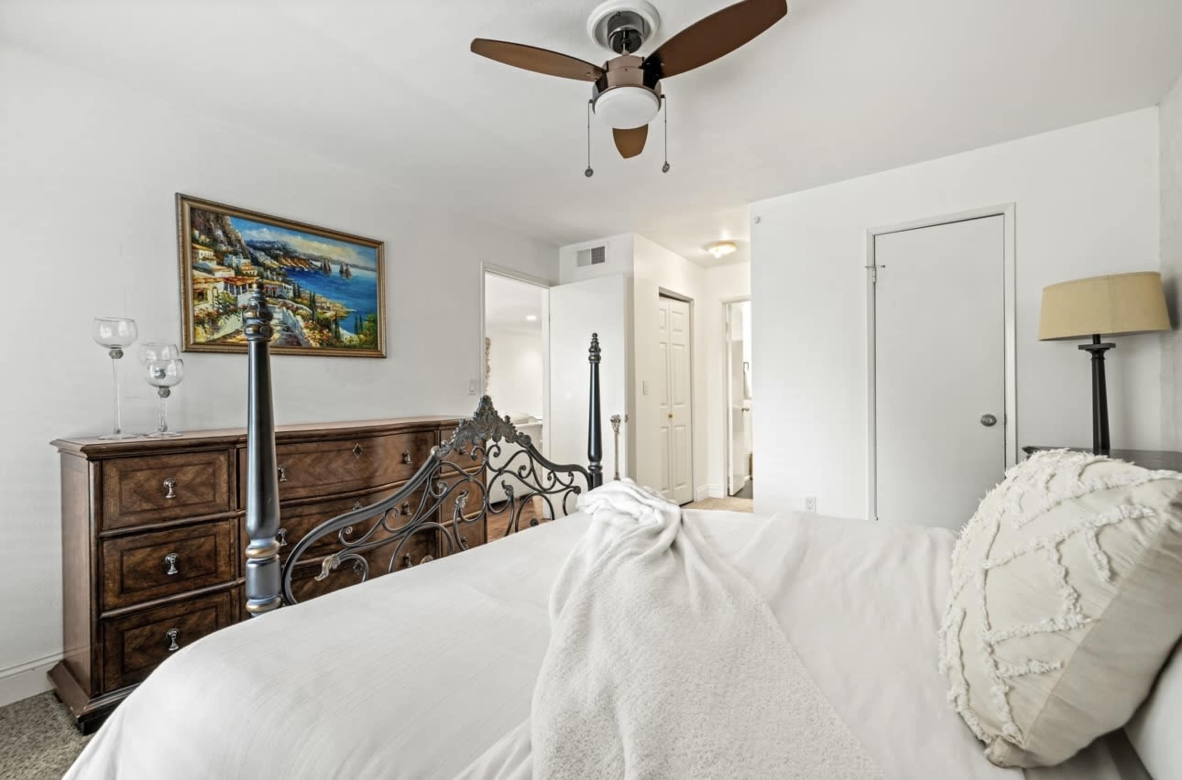 The image shows a neatly arranged bedroom featuring a wooden dresser, a bed with a decorative headboard, and a ceiling fan.