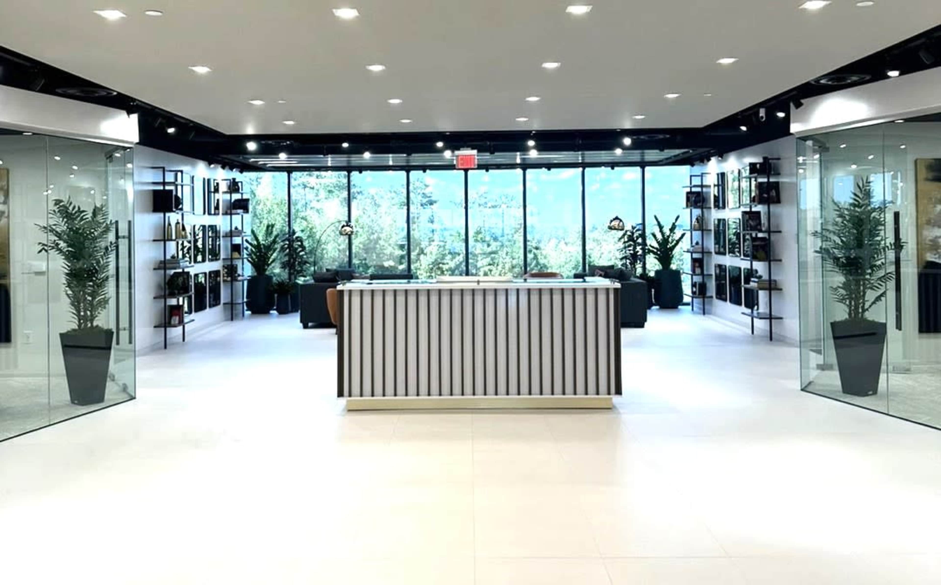 The image shows a modern reception area with a striped front desk, surrounded by glass walls and potted plants, leading to a view of an outdoor landscape.