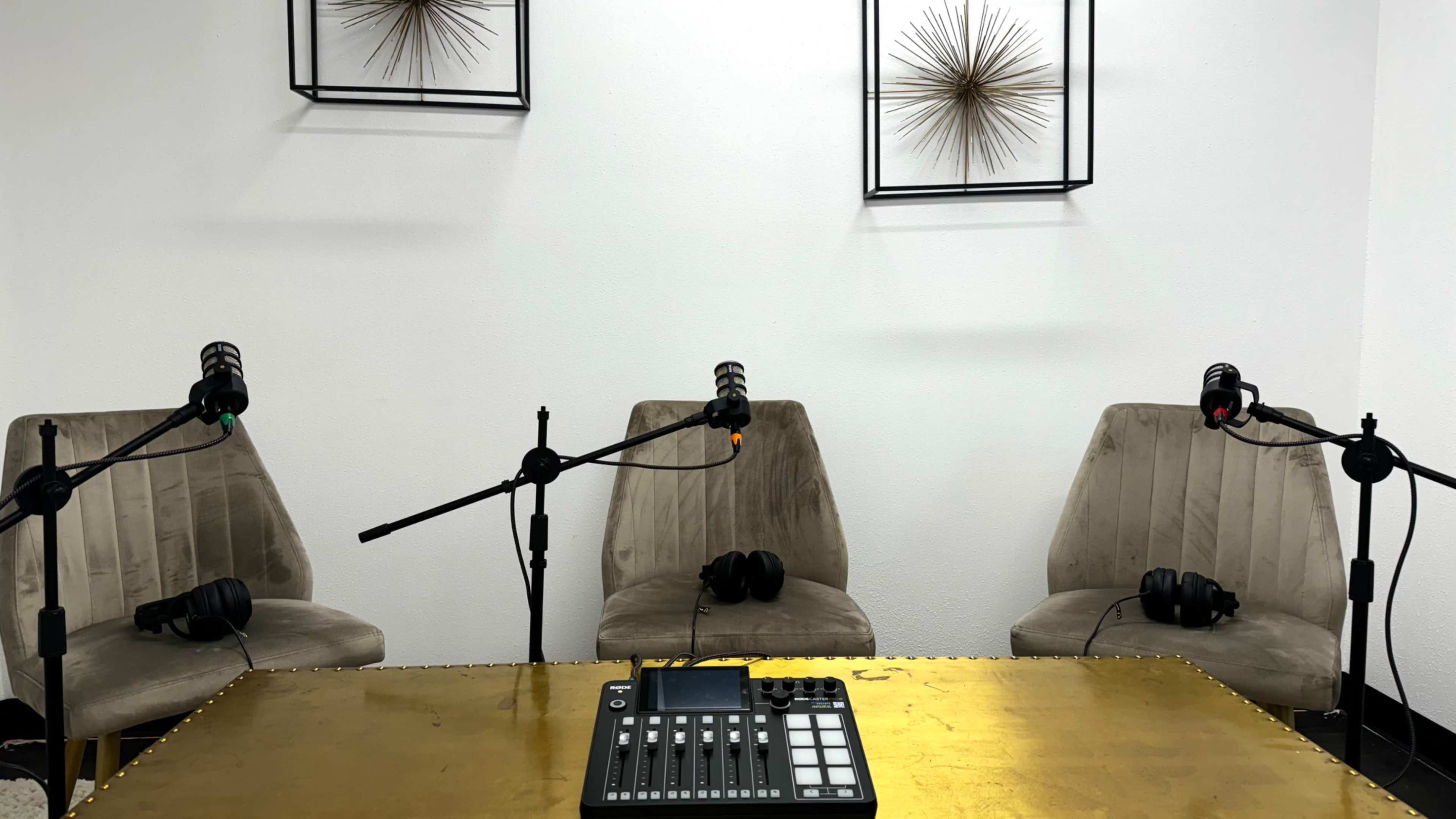 The image shows a small, modern recording studio setup with three microphones positioned over empty chairs, a table with a sound mixer, and decorative wall art in the background.