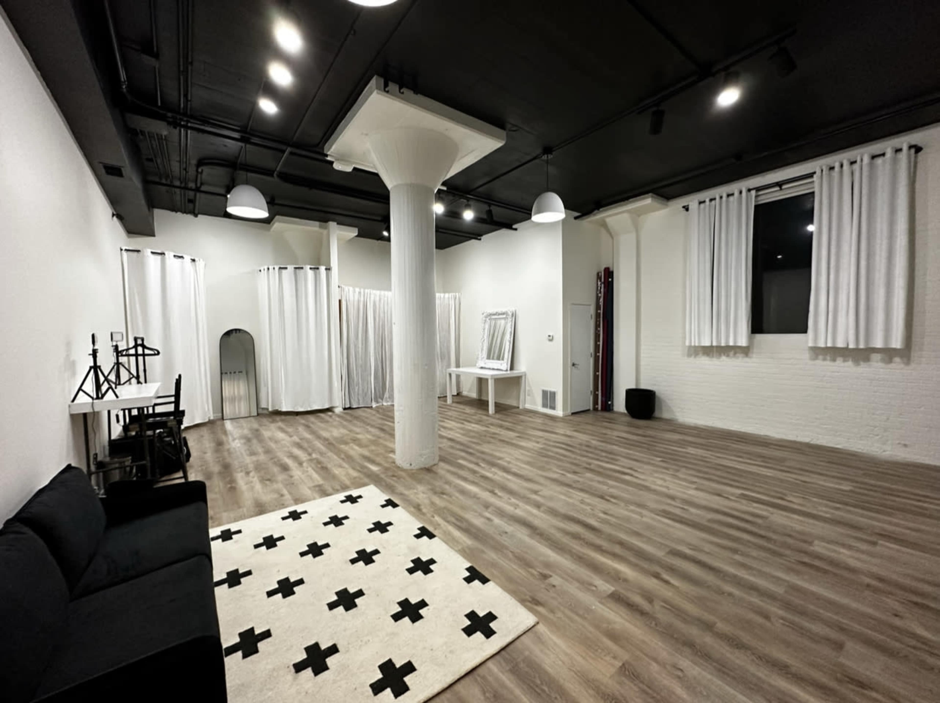 A spacious, empty room with wooden flooring, a white wall, a sofa, and a large area rug featuring a black cross pattern.