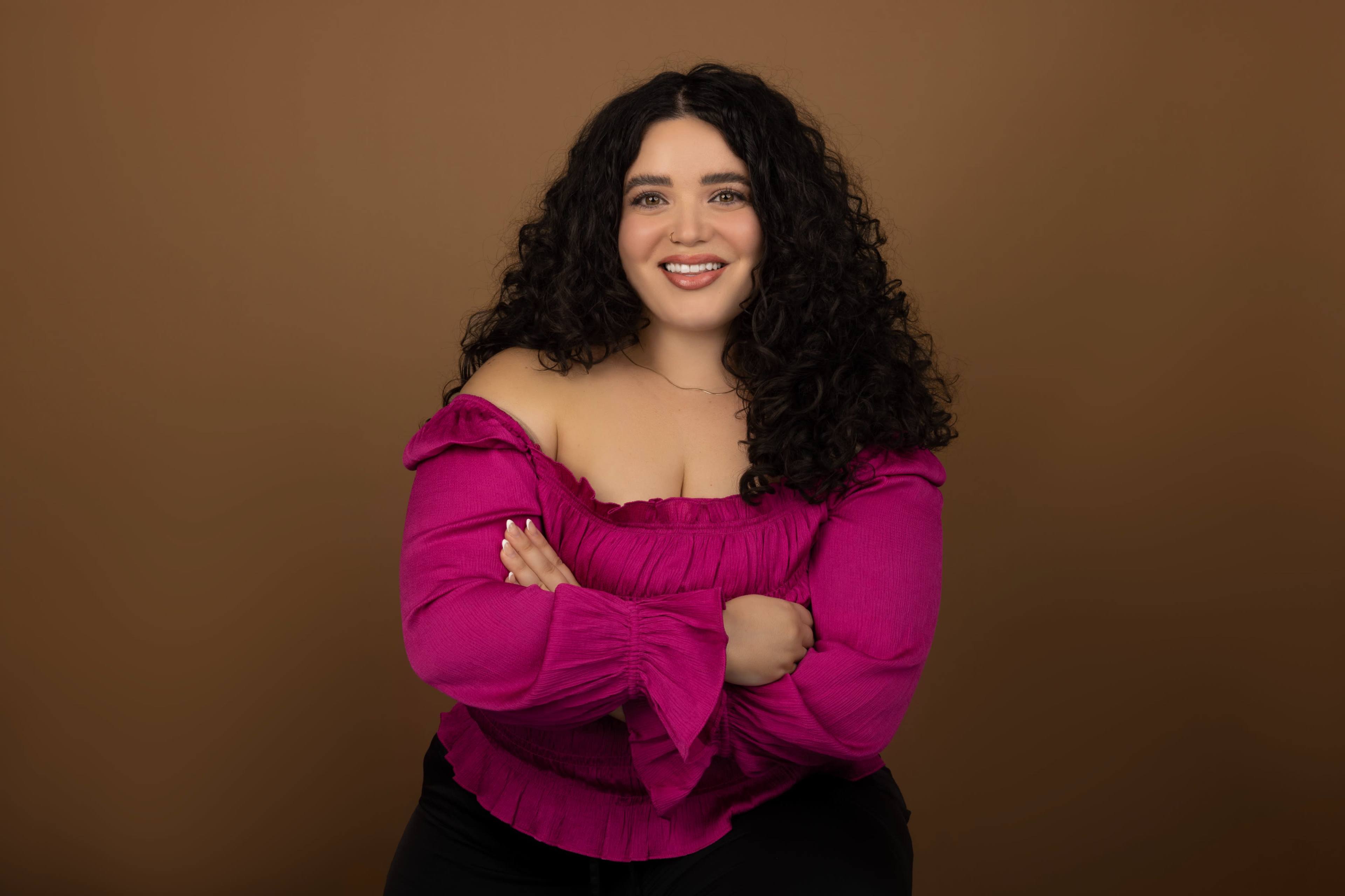 A woman with curly hair is sitting and smiling, wearing a pink off-shoulder top against a brown background.