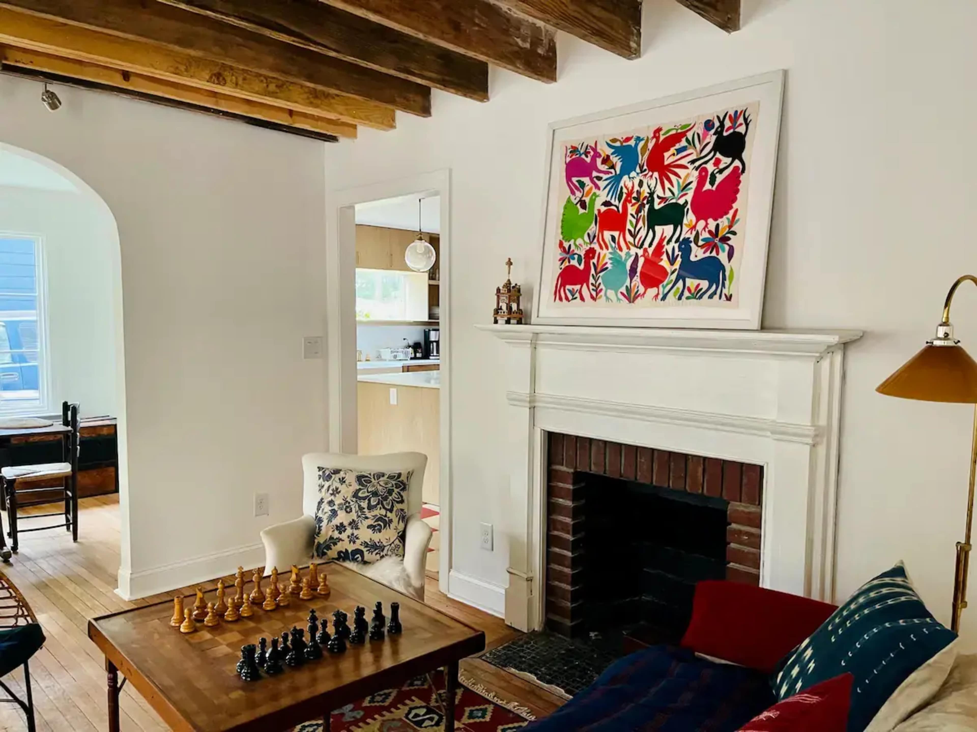 A living room with a chess set on a wooden table, a colorful artwork above the fireplace, and light-colored wooden beams on the ceiling.