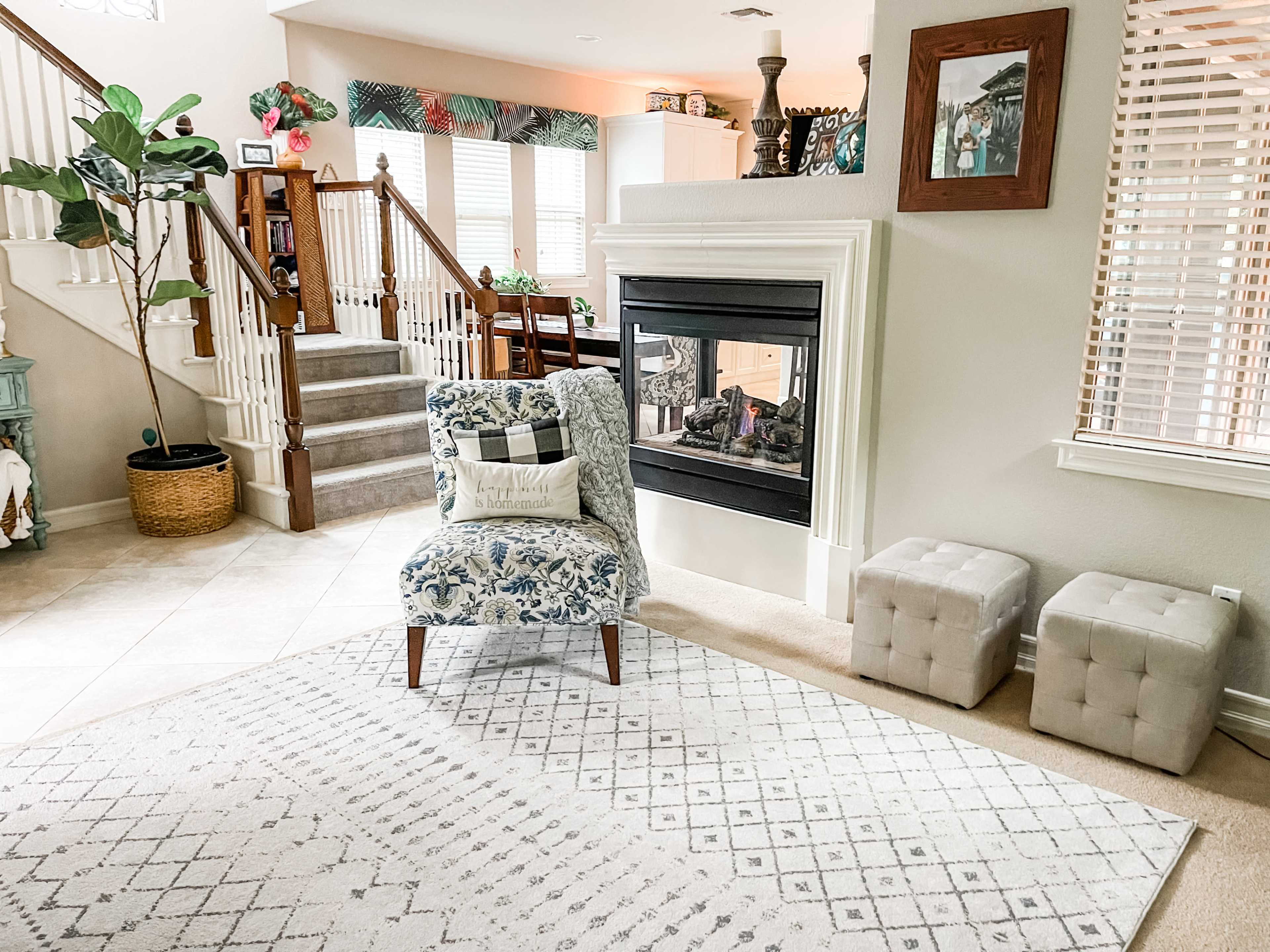 A living room with a patterned armchair near a fireplace, surrounded by a staircase, a dining area, and decorative elements.