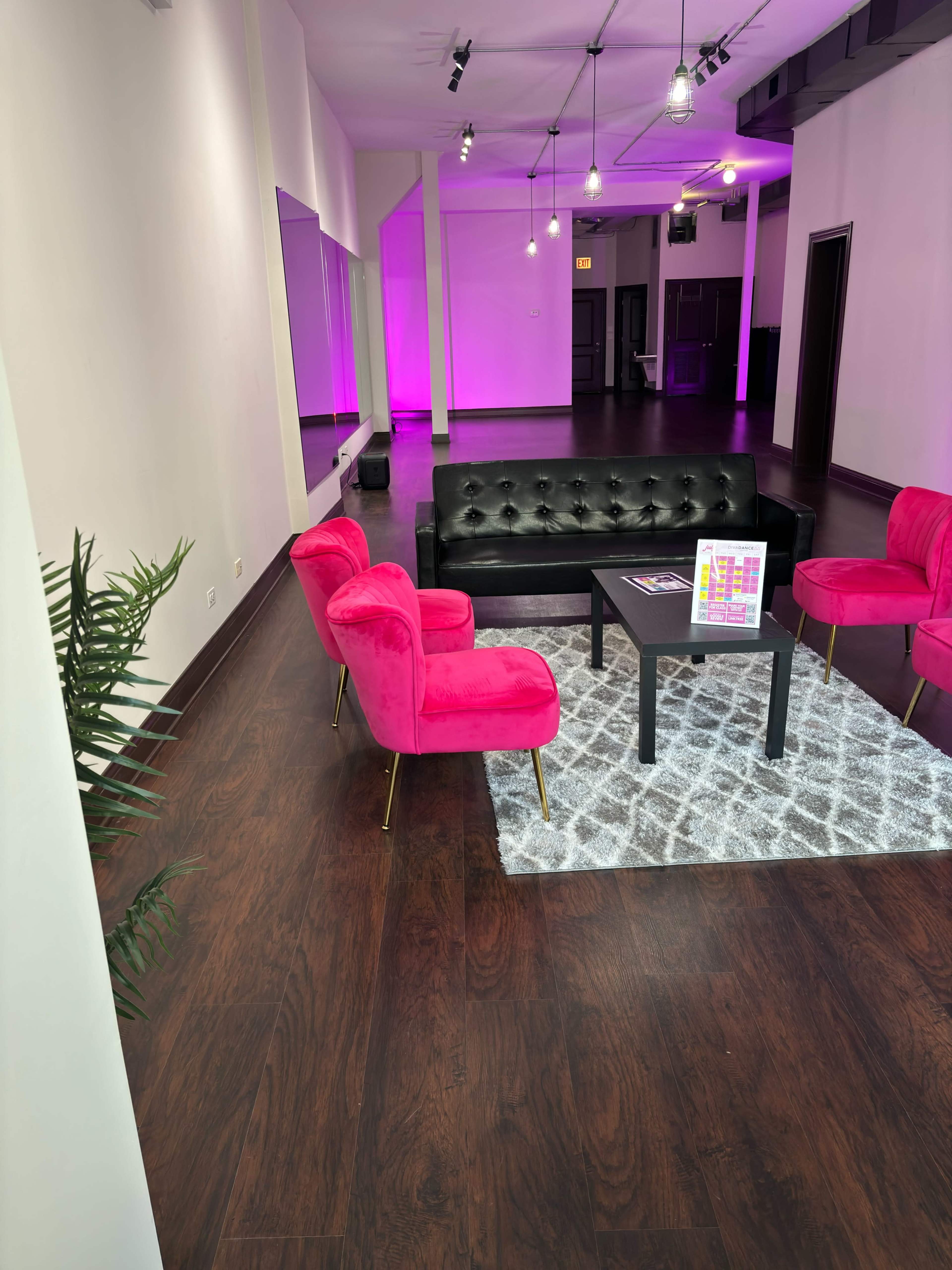 A modern lounge area with a black leather sofa, pink chairs, a small black table on a patterned rug, and soft purple lighting.