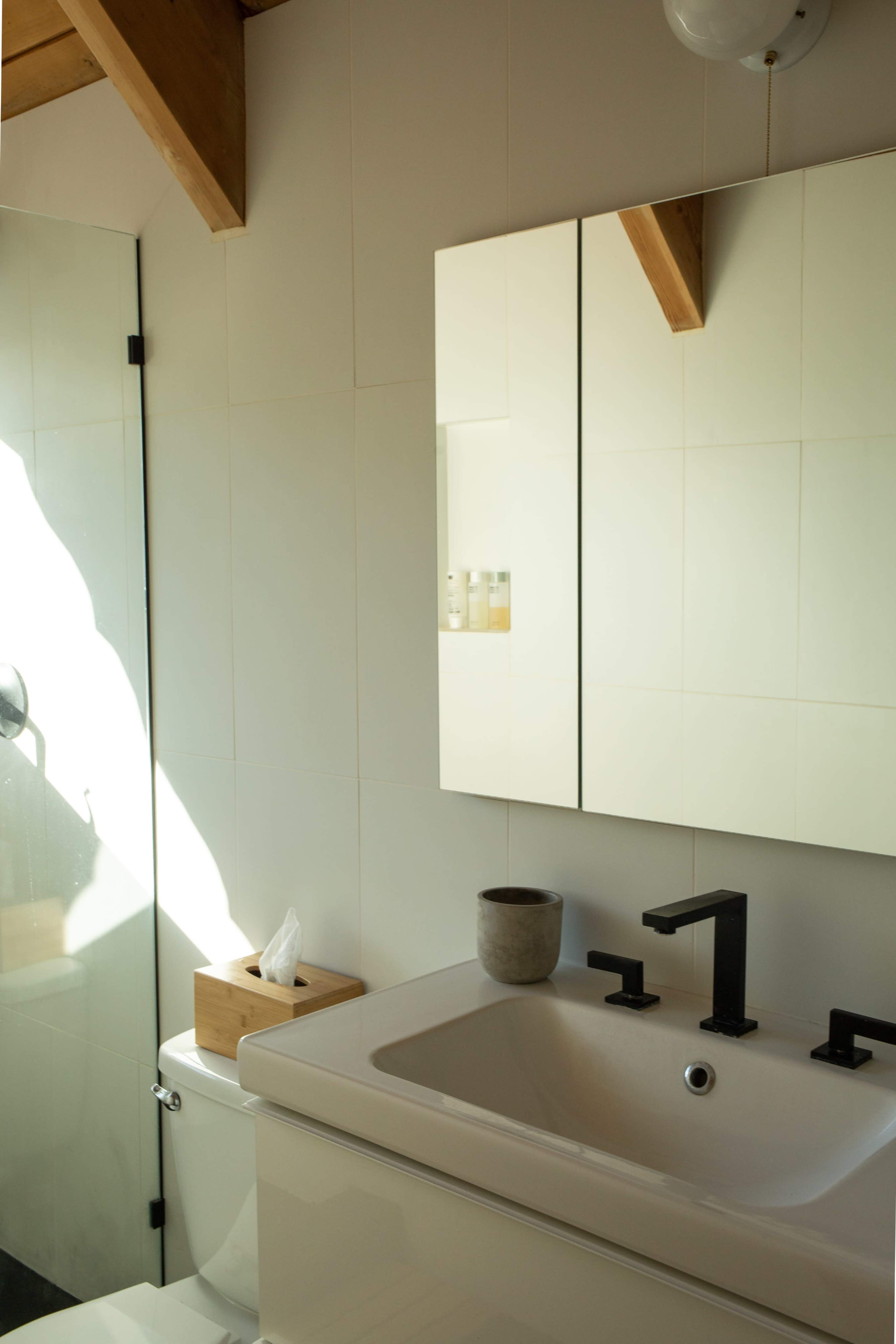A modern bathroom with a white sink and a large mirror, accompanied by a glass shower, wooden accents, and a tissue box.