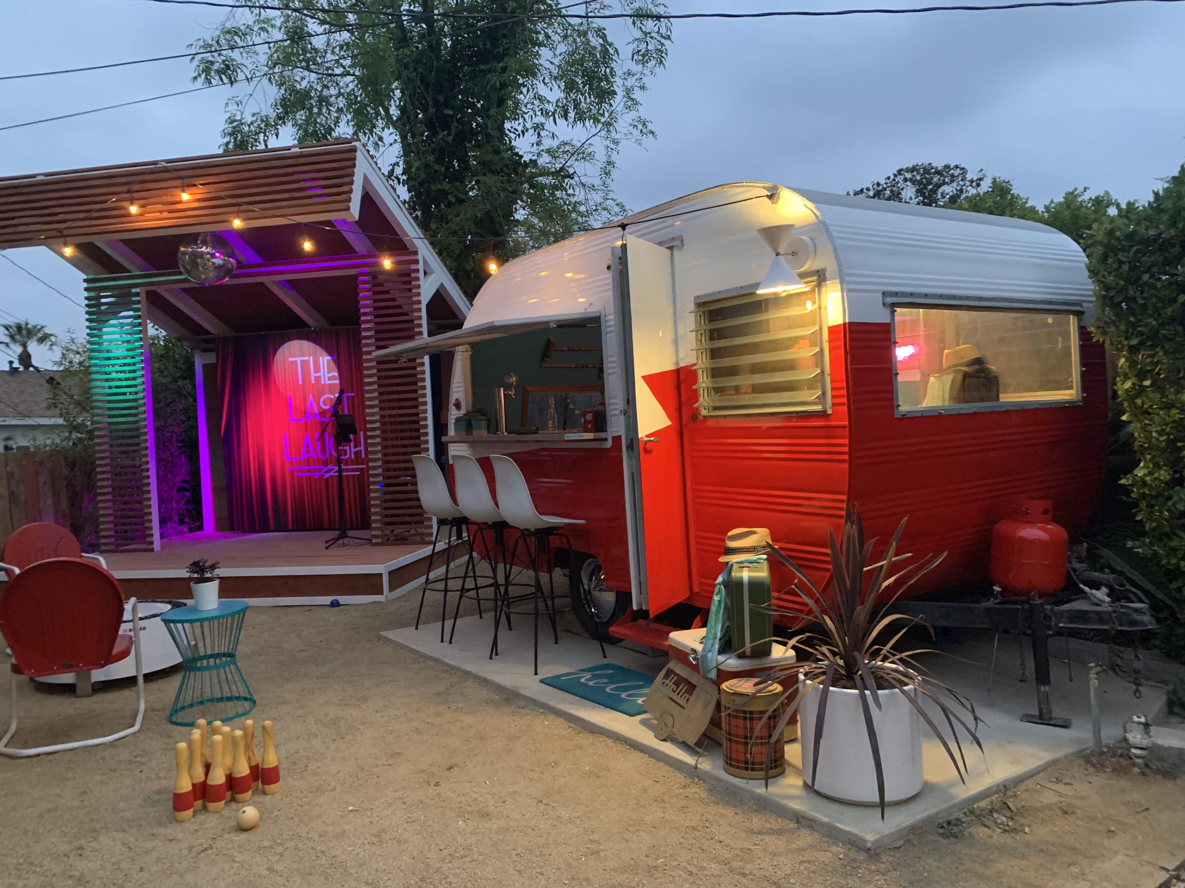 A vintage red and white camper is parked beside a small outdoor stage with colorful lighting and wooden structures in a backyard setting.