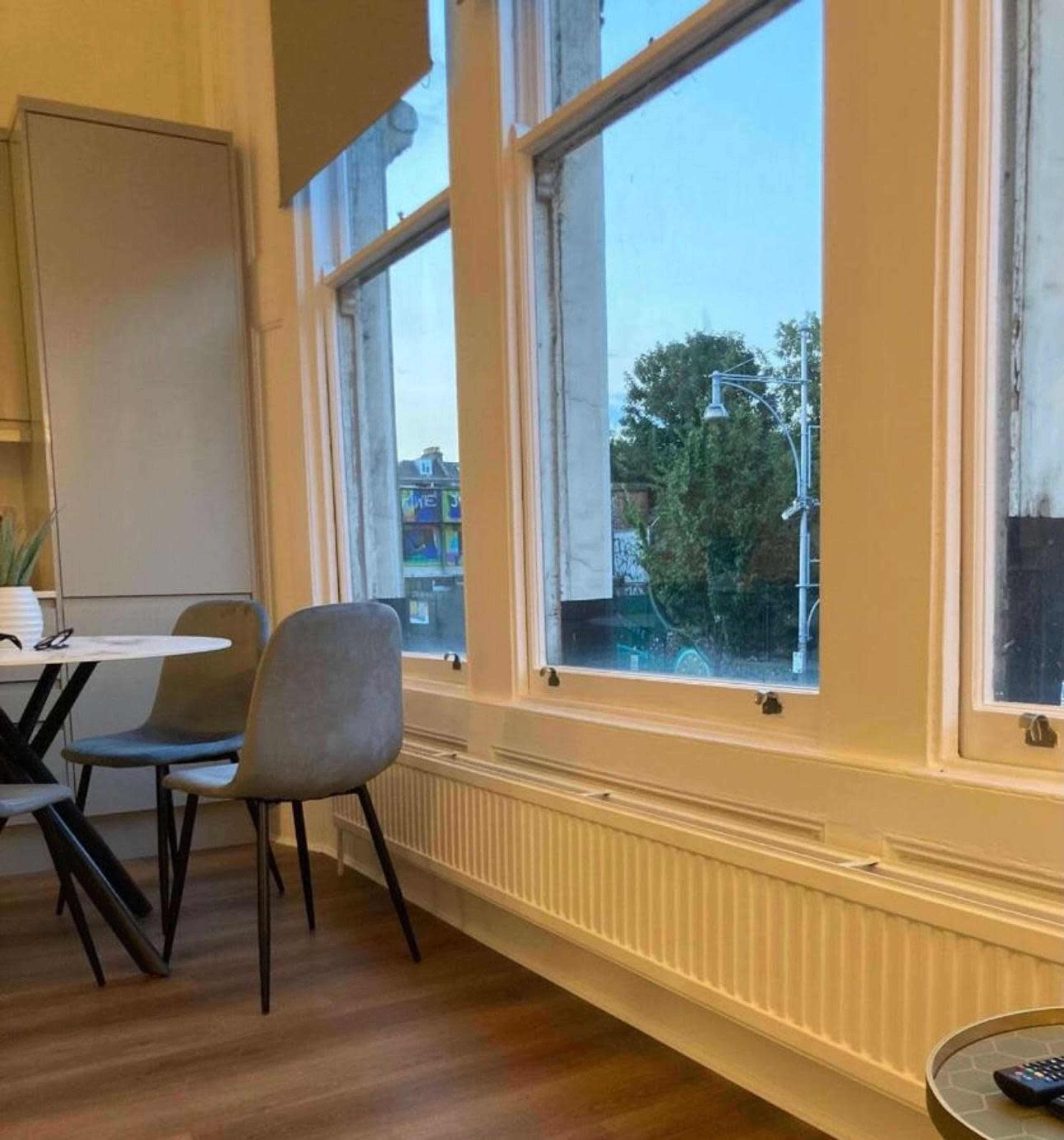 The image shows a corner of a room with a round dining table and chairs, featuring large windows that overlook an outdoor view.
