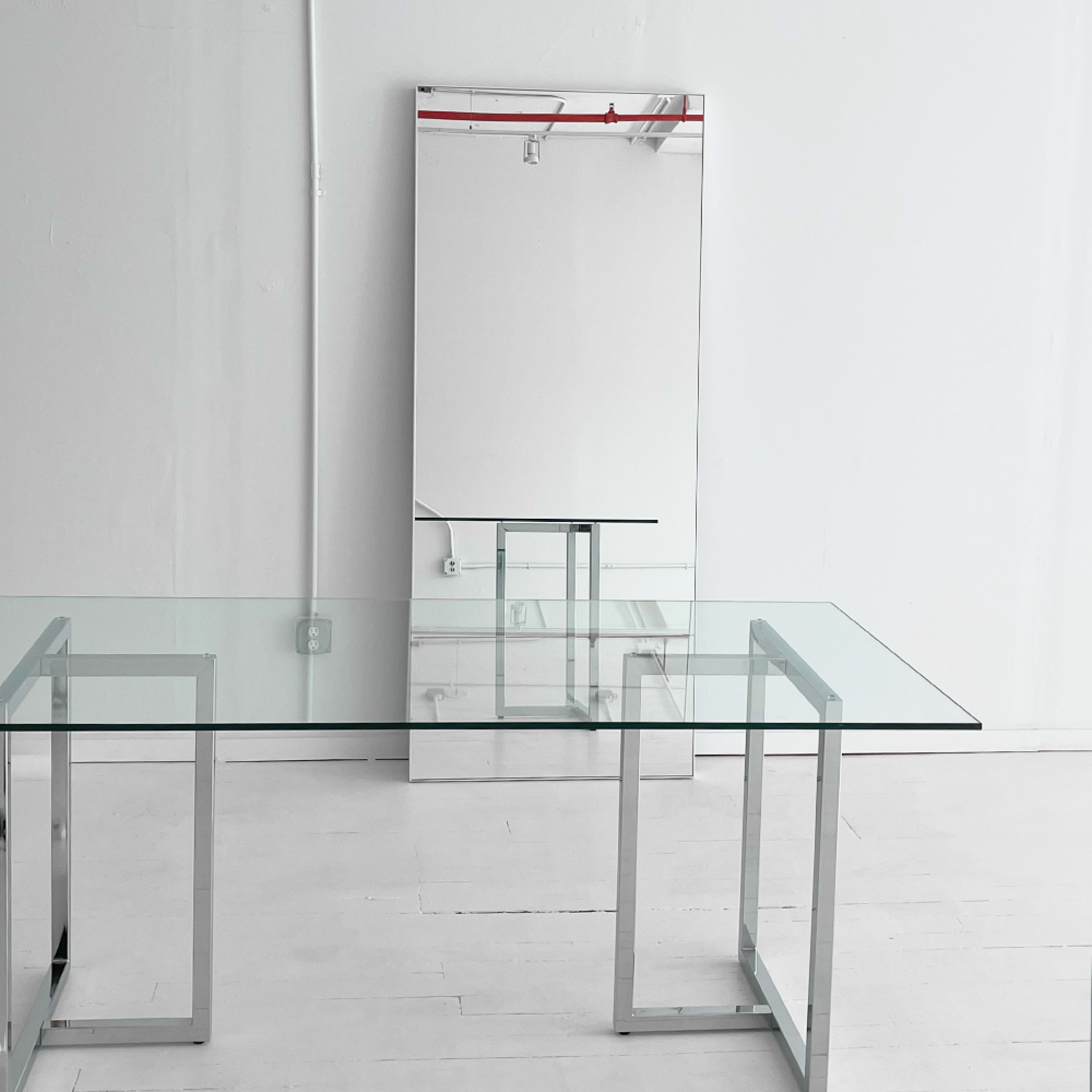 A clear glass table with a metal frame stands next to a large rectangular mirror in a minimalistic white room.