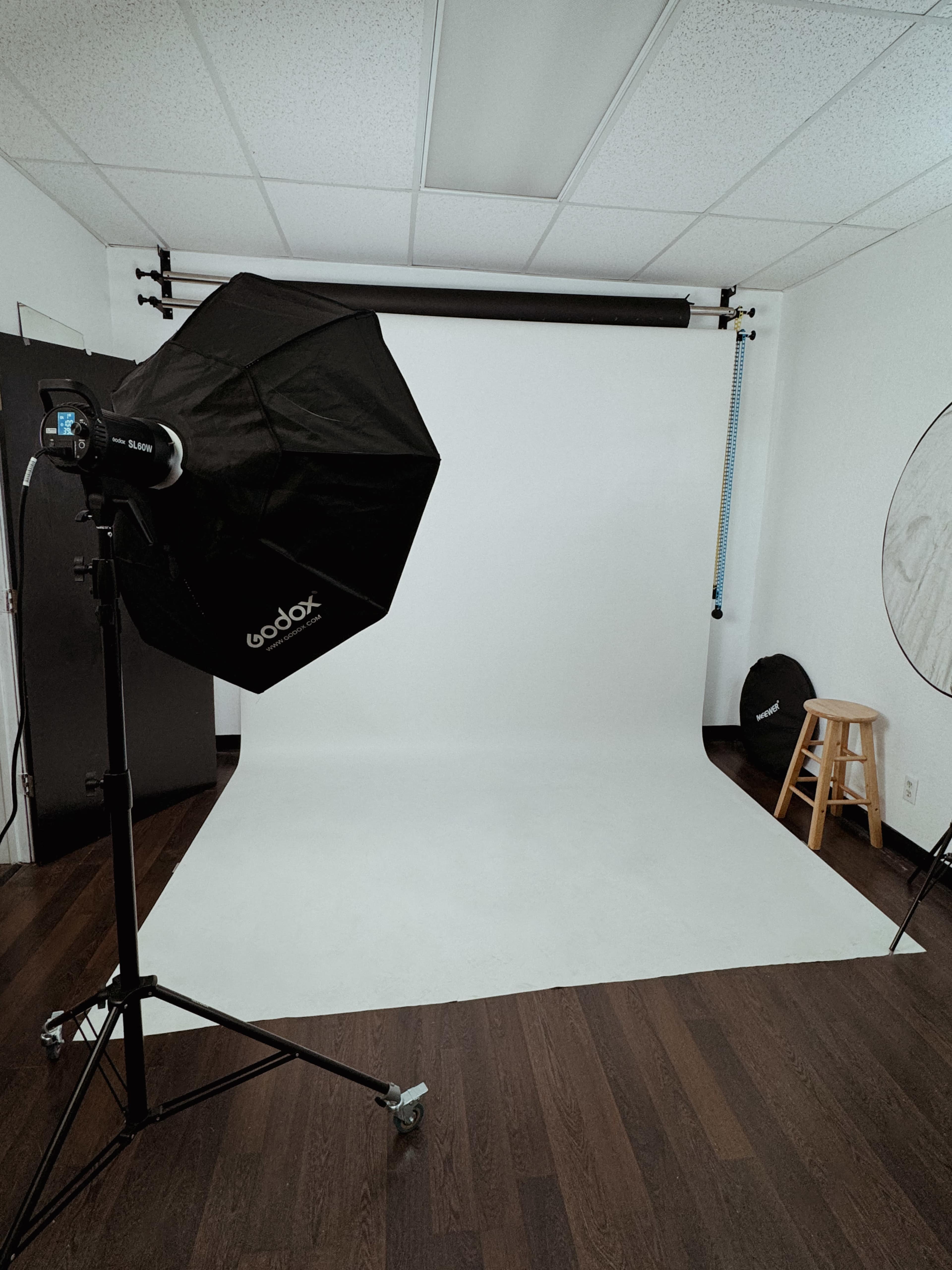 The image shows a photography studio setup with a softbox light, a white backdrop, and a stool against a wooden floor.