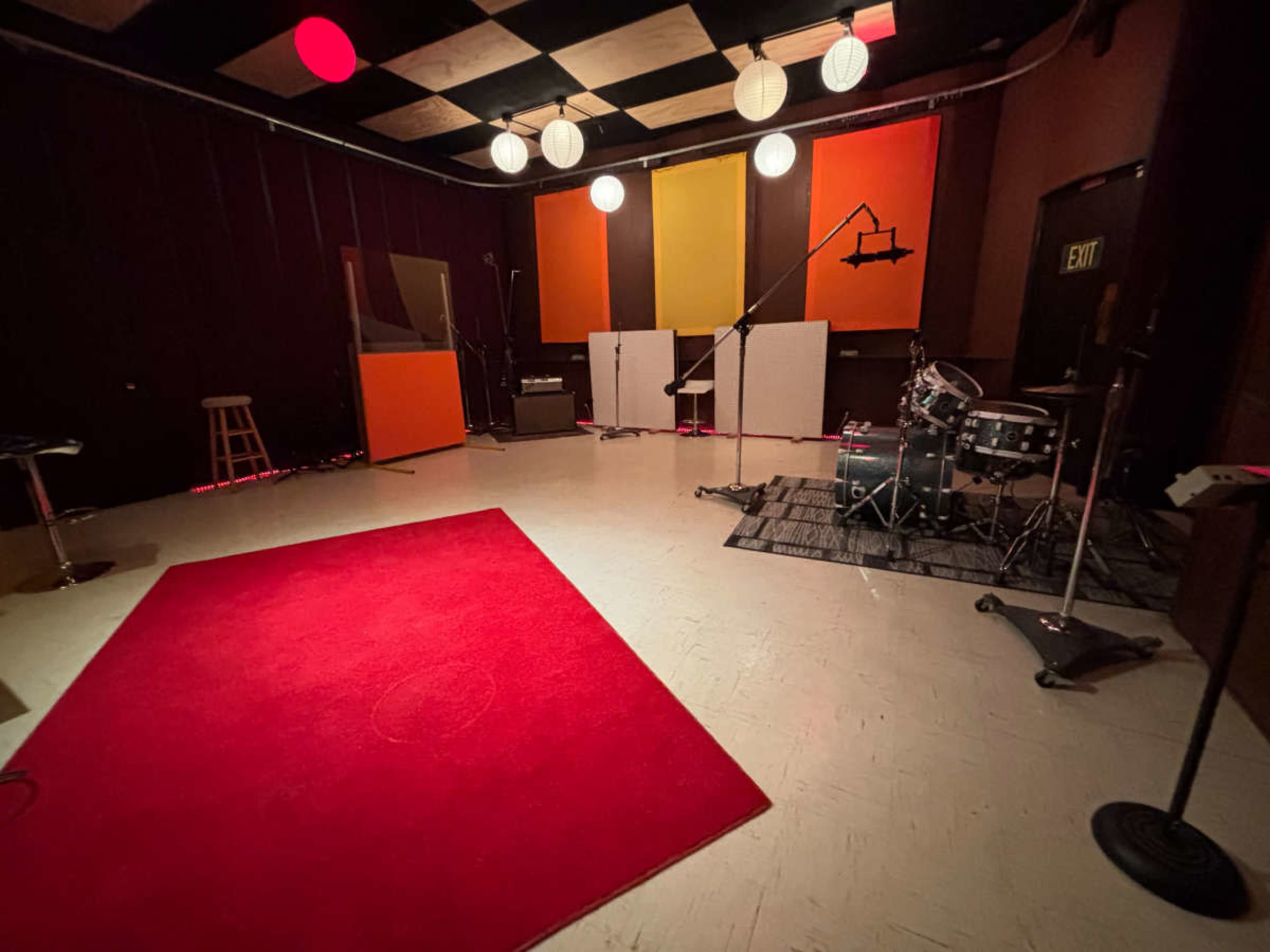 The image shows a dimly lit music studio featuring a red area rug, a small drum set, amplifiers, and colorful acoustic panels on the walls.