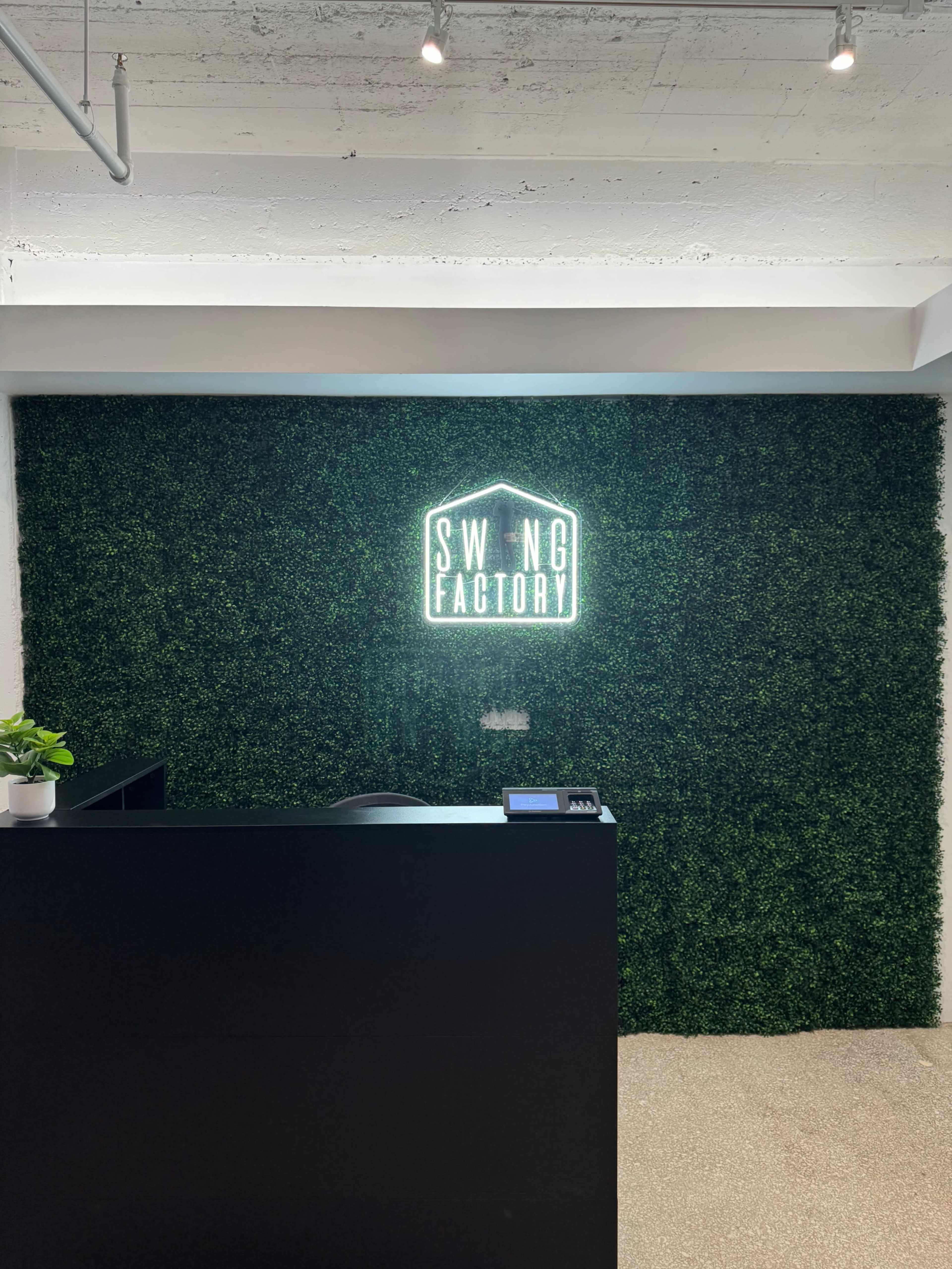 A reception area with a green hedge wall and a illuminated sign that reads "SWING FACTORY."