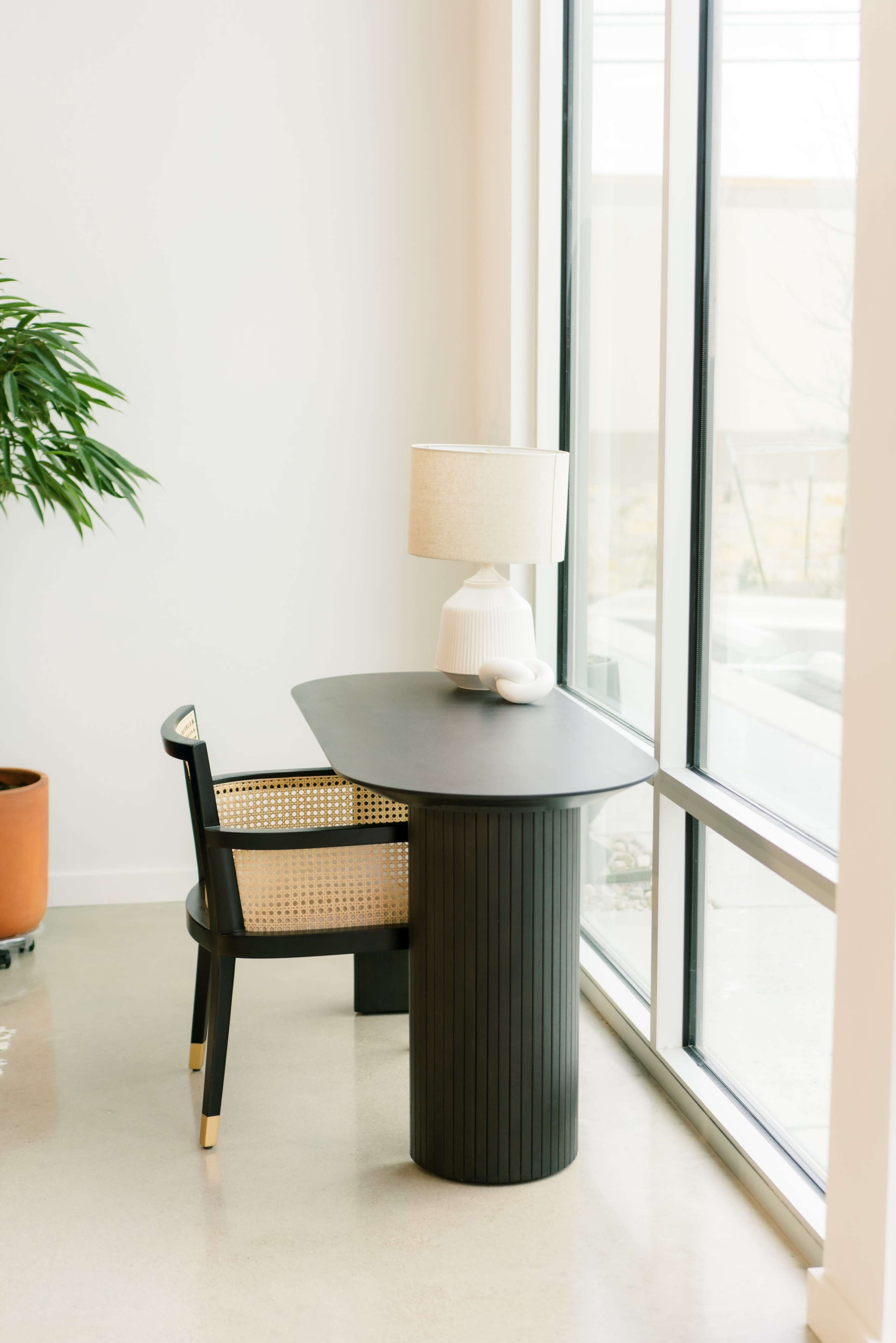 A black desk with a lamp and a chair is positioned near large windows in a well-lit room.
