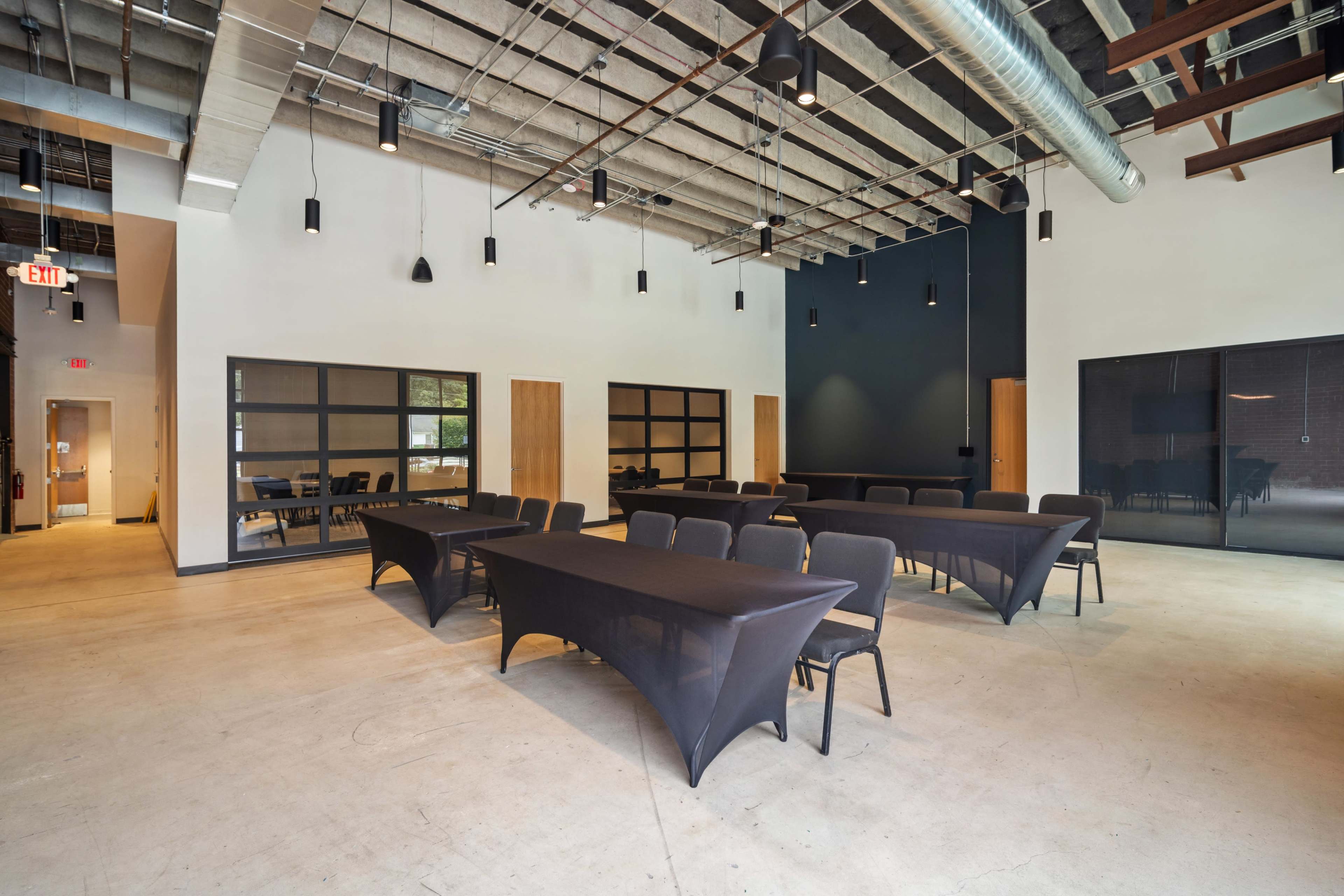 The image shows a spacious, modern conference room with black tables arranged in the center and chairs facing them, illuminated by pendant lights.