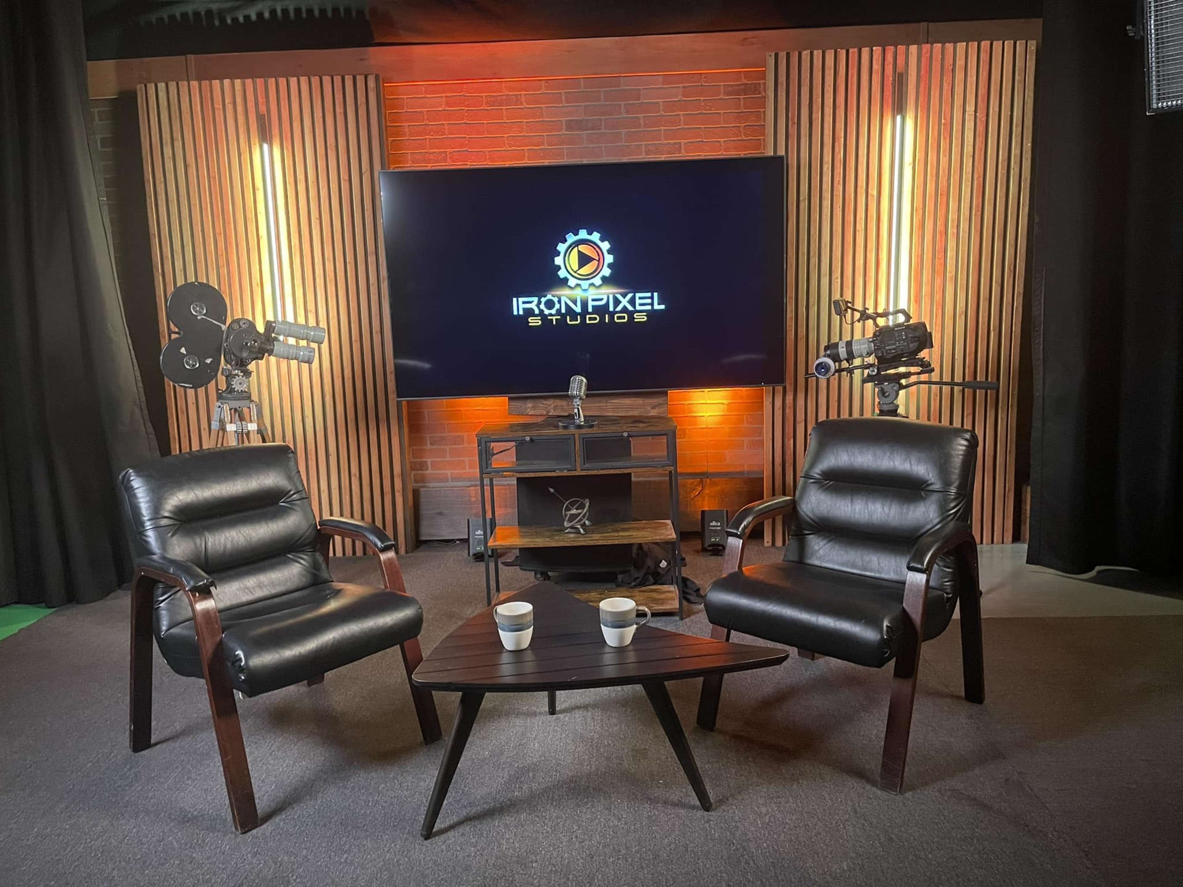 The image shows a modern studio setting with two black leather chairs facing a wooden table, a television displaying the "IRONPIXEL STUDIOS" logo, and film cameras mounted on either side.